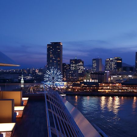 神戸メリケンパークオリエンタルホテル 神戸市 エクステリア 写真 The photo displays a panoramic view of a waterfront cityscape at twilight. You can see a vibrant skyline with towering buildings lit up against the evening sky. In the foreground, there are modern architectural elements, possibly part of a hotel or r