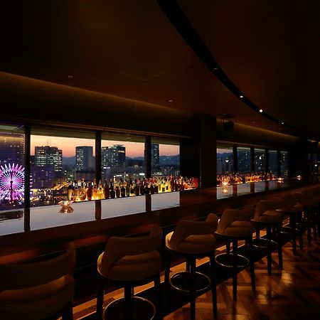 神戸メリケンパークオリエンタルホテル 神戸市 エクステリア 写真 The photo shows the interior of a stylish bar or lounge with a panoramic view of a city skyline at dusk. The lighting is dim, creating an intimate atmosphere. There are elegant bar stools lined up along a long counter, which features an array of illu
