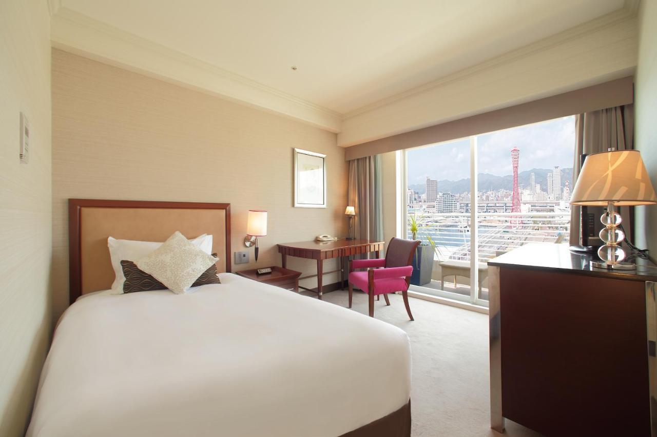 神戸メリケンパークオリエンタルホテル 神戸市 エクステリア 写真 The photo shows a hotel room featuring a single bed with white bedding and decorative pillows. There is a wooden desk and chair positioned near a large window that offers a view of the outside, likely a cityscape or waterfront. A small pink chair add