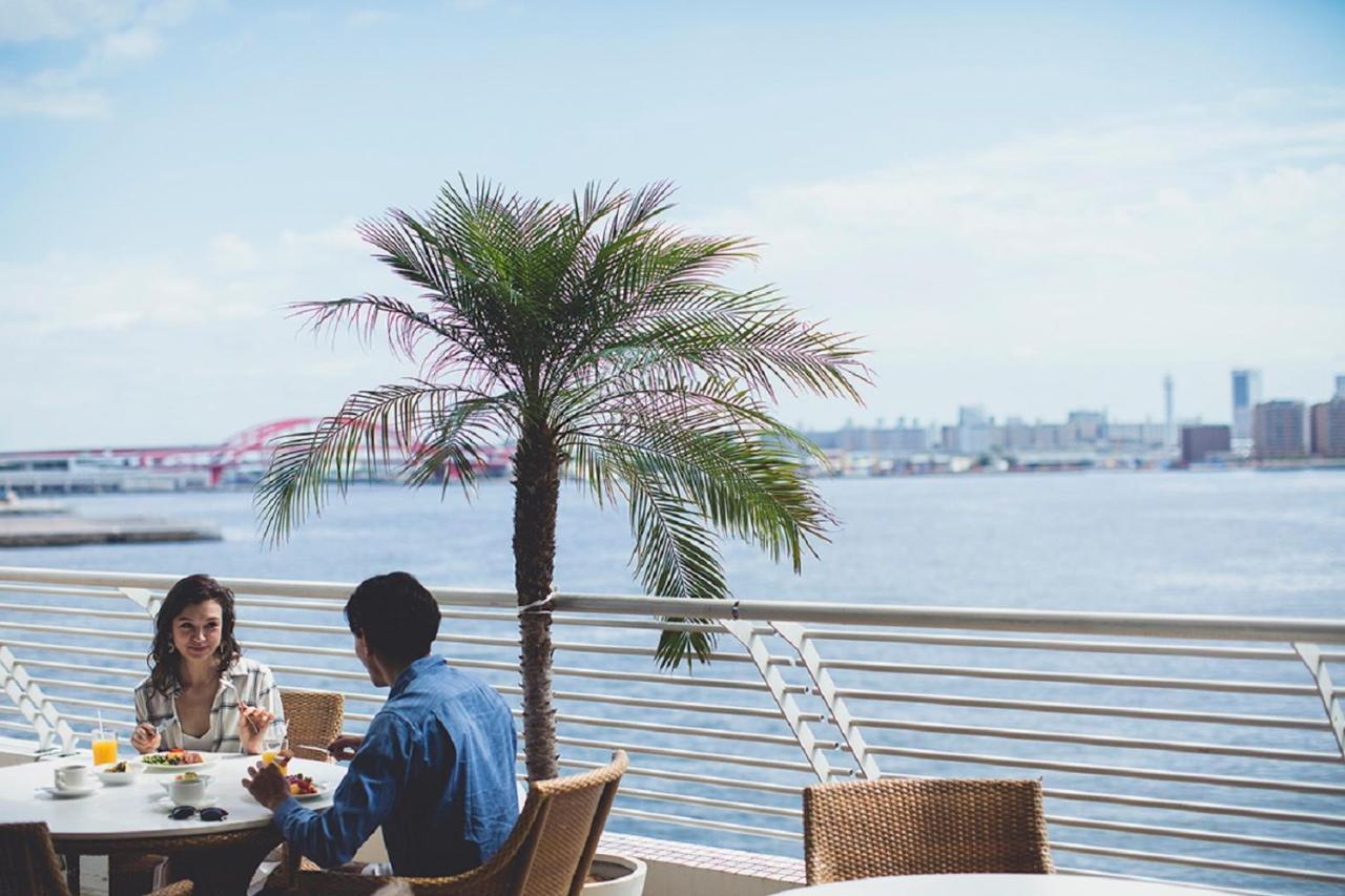 神戸メリケンパークオリエンタルホテル 神戸市 エクステリア 写真 The photo depicts a scenic outdoor dining setting by the water. In the foreground, there are two people sitting at a table with a stylish arrangement of food and drinks. A palm tree provides a tropical ambiance, standing prominently next to them. In 