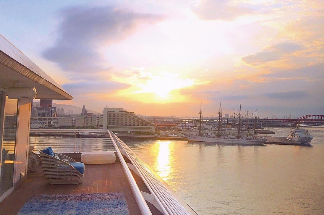 神戸メリケンパークオリエンタルホテル 神戸市 エクステリア 写真 The photo shows a serene waterfront scene with a stunning sunset. In the foreground, there is a deck area that appears to be part of a ship or a waterfront building, featuring a cozy seating arrangement. The deck has a soft rug and some cushions, sug