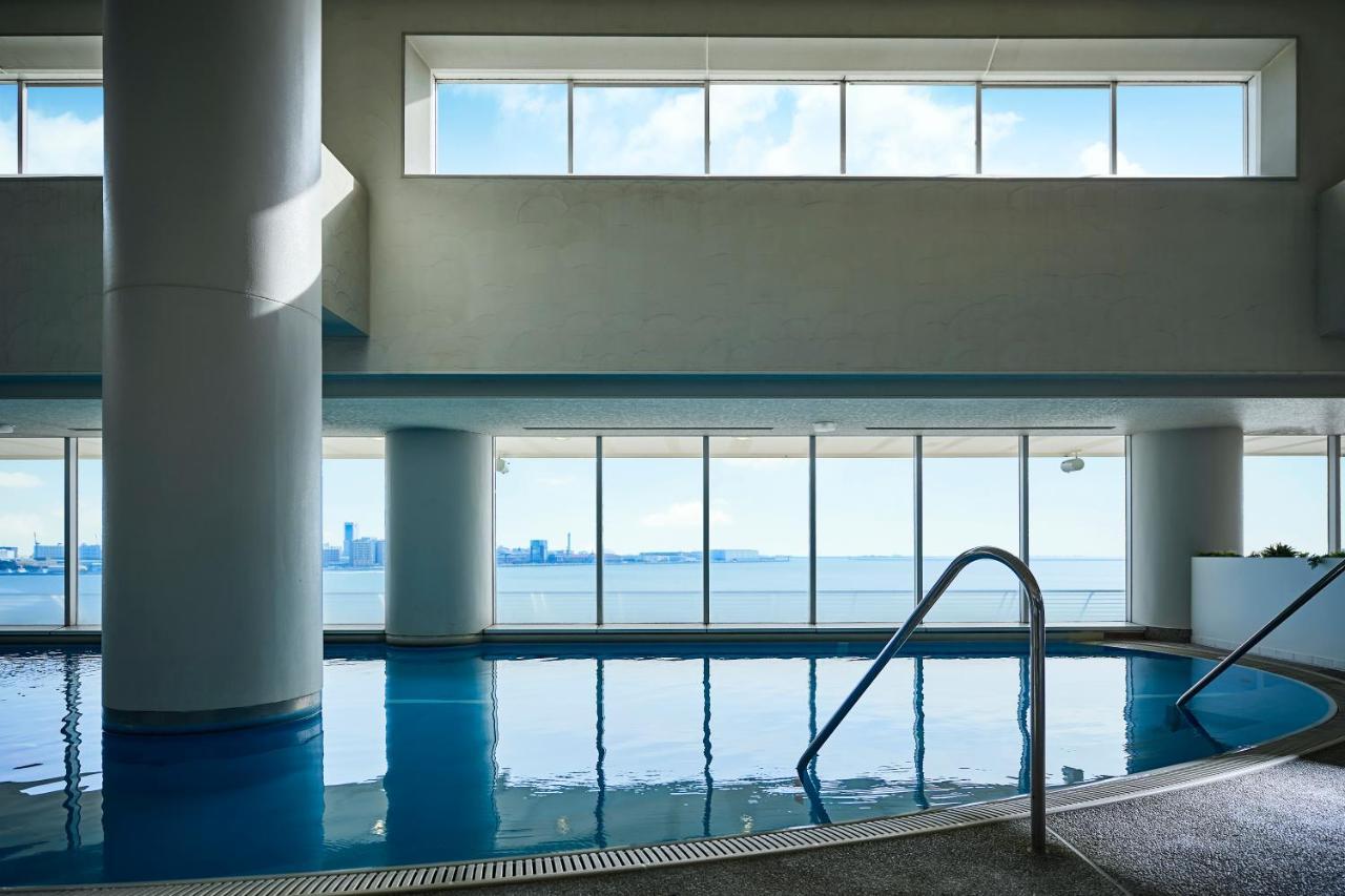 神戸メリケンパークオリエンタルホテル 神戸市 エクステリア 写真 The photo shows an indoor swimming pool with a clear view through large windows. The pool has smooth, reflective water and a ladder leading into it. In the background, there are distant buildings and a waterfront visible through the windows, suggesti