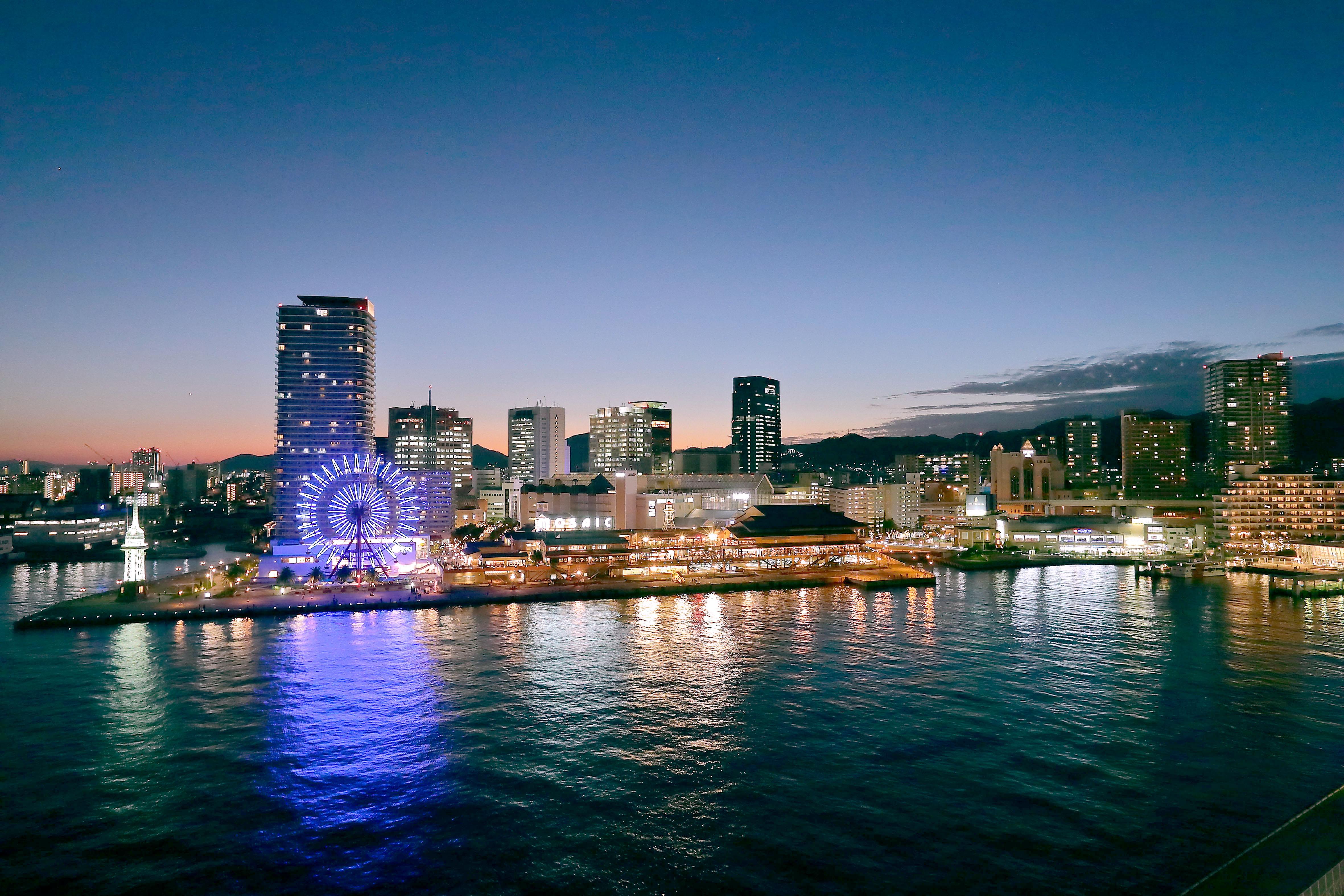 神戸メリケンパークオリエンタルホテル 神戸市 エクステリア 写真 The photo depicts a cityscape at dusk, showcasing a waterfront area. In the foreground, the water reflects the fading light, creating a tranquil scene. On the shore, there are several tall buildings illuminated by city lights, contributing to the urb