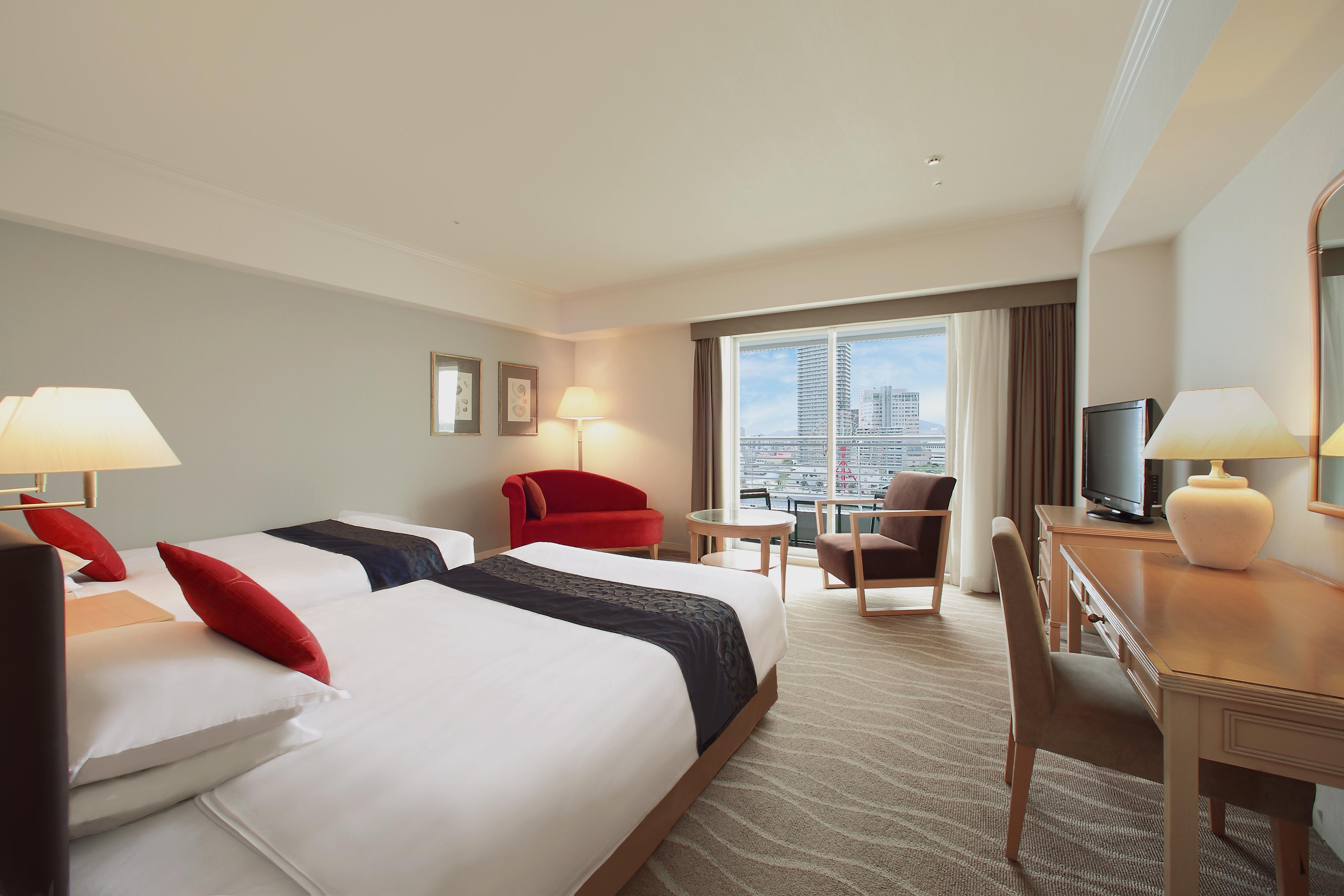 神戸メリケンパークオリエンタルホテル 神戸市 エクステリア 写真 The photo shows a hotel room featuring two single beds with white linens and red accent pillows. There is a red chair near a window with a view, and a small wooden desk with a chair next to it. A television is positioned on a shelf, and there are lam