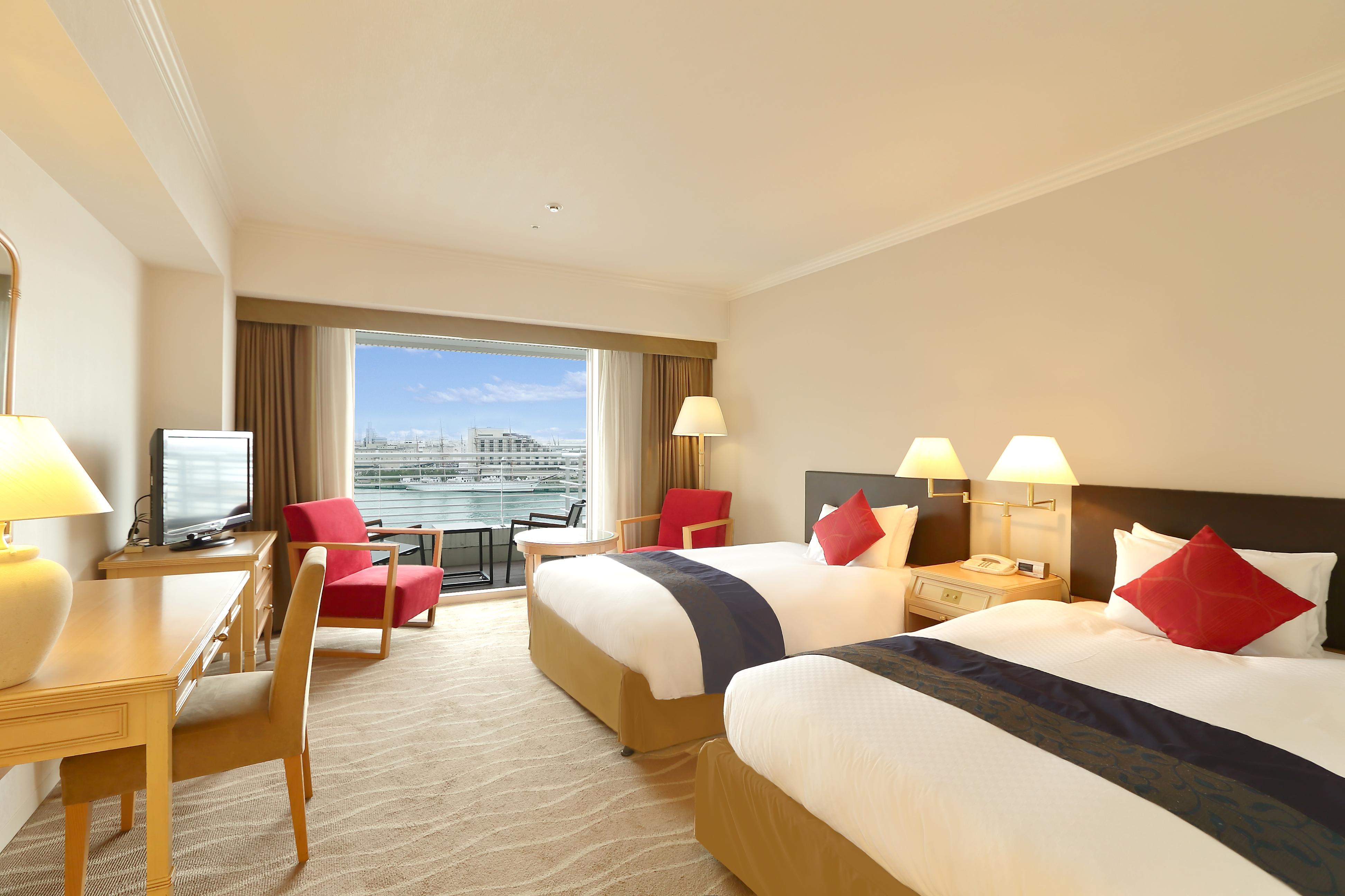 神戸メリケンパークオリエンタルホテル 神戸市 エクステリア 写真 The photo shows a hotel room with two beds, each adorned with white linens and decorative red pillows. There is a television mounted on the wall and a wooden desk with a chair on one side of the room. Two comfortable red armchairs are positioned near