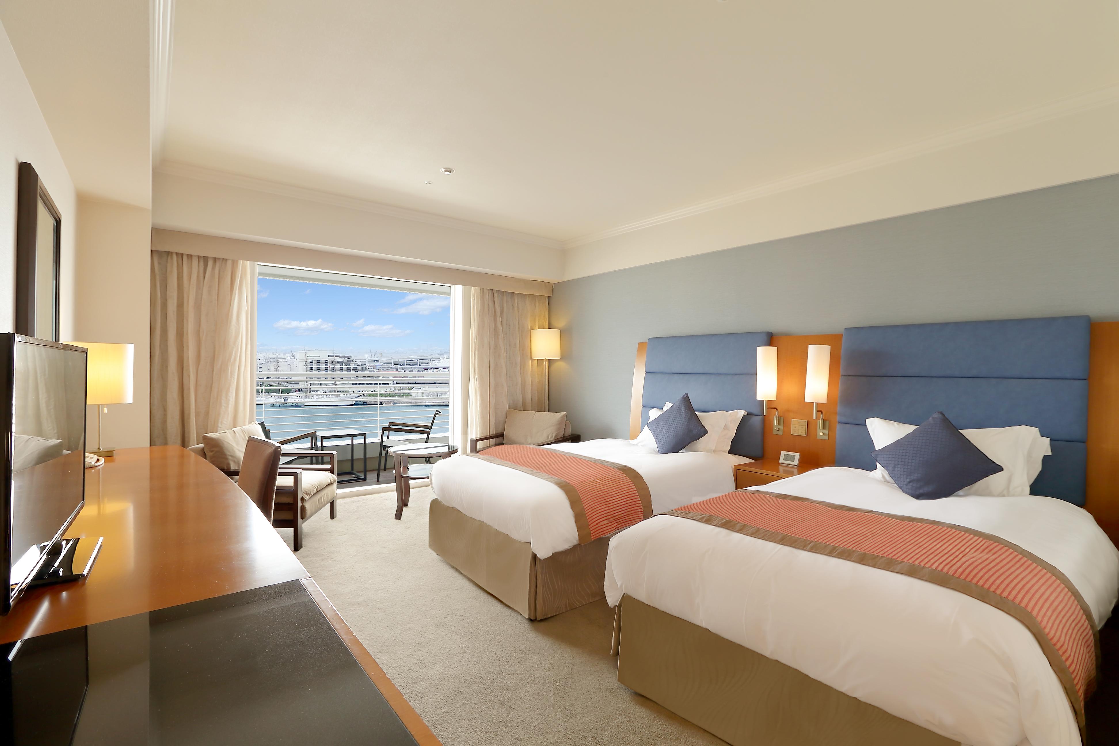 神戸メリケンパークオリエンタルホテル 神戸市 エクステリア 写真 The photo shows a hotel room that features two beds with white linens and orange accents. There is a wooden desk and chair positioned near the entrance, along with a modern chair and a small table next to the window. The room has large windows that a