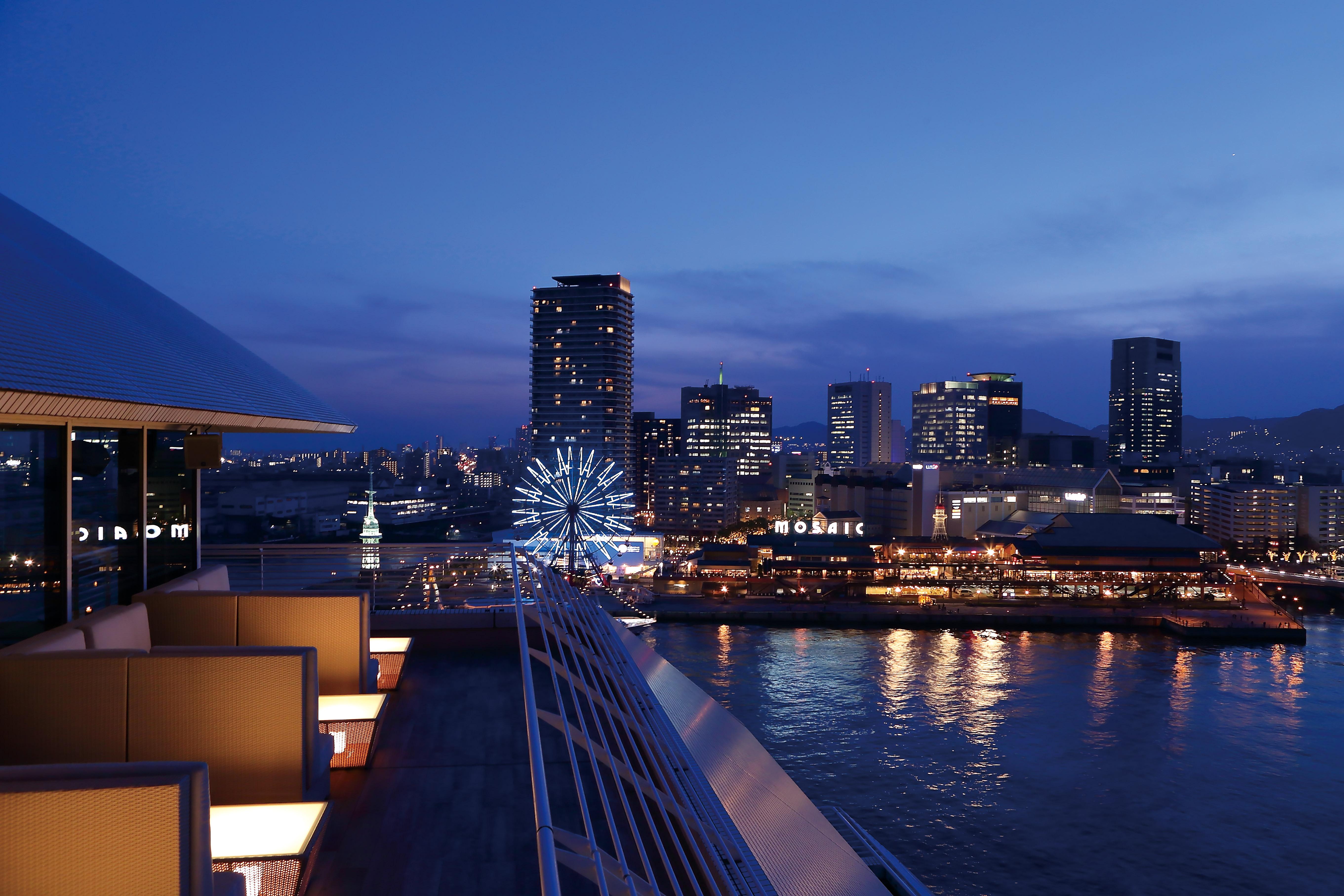 神戸メリケンパークオリエンタルホテル 神戸市 エクステリア 写真 The photo displays a panoramic view of a waterfront cityscape at twilight. You can see a vibrant skyline with towering buildings lit up against the evening sky. In the foreground, there are modern architectural elements, possibly part of a hotel or r