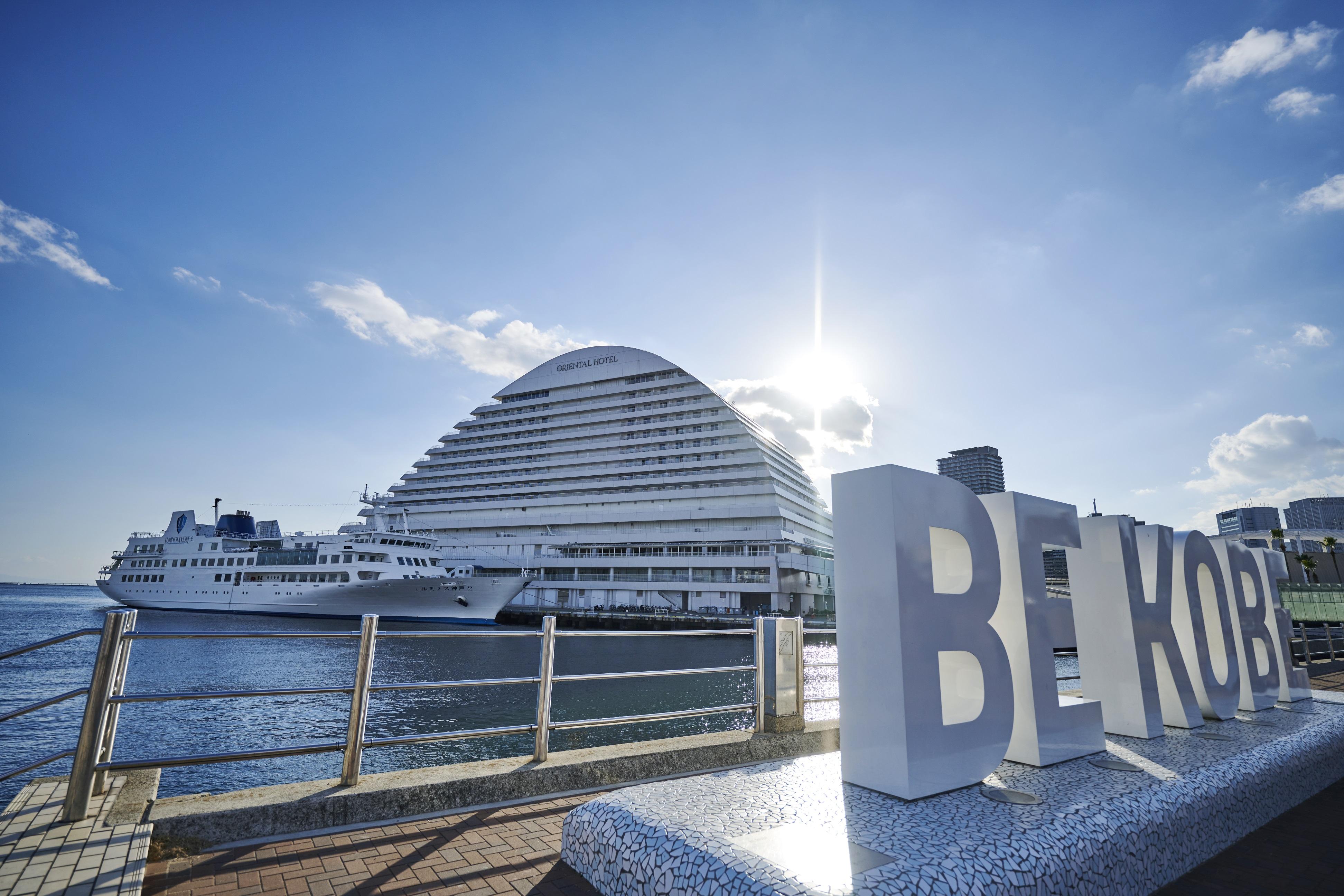 神戸メリケンパークオリエンタルホテル 神戸市 エクステリア 写真 The photo depicts a waterfront scene in Kobe, Japan. In the foreground, there is a large sign that reads "BE KOBE." Behind the sign, a modern, uniquely shaped building resembling a ship is visible, along with a cruise liner docked in the harbor. The 
