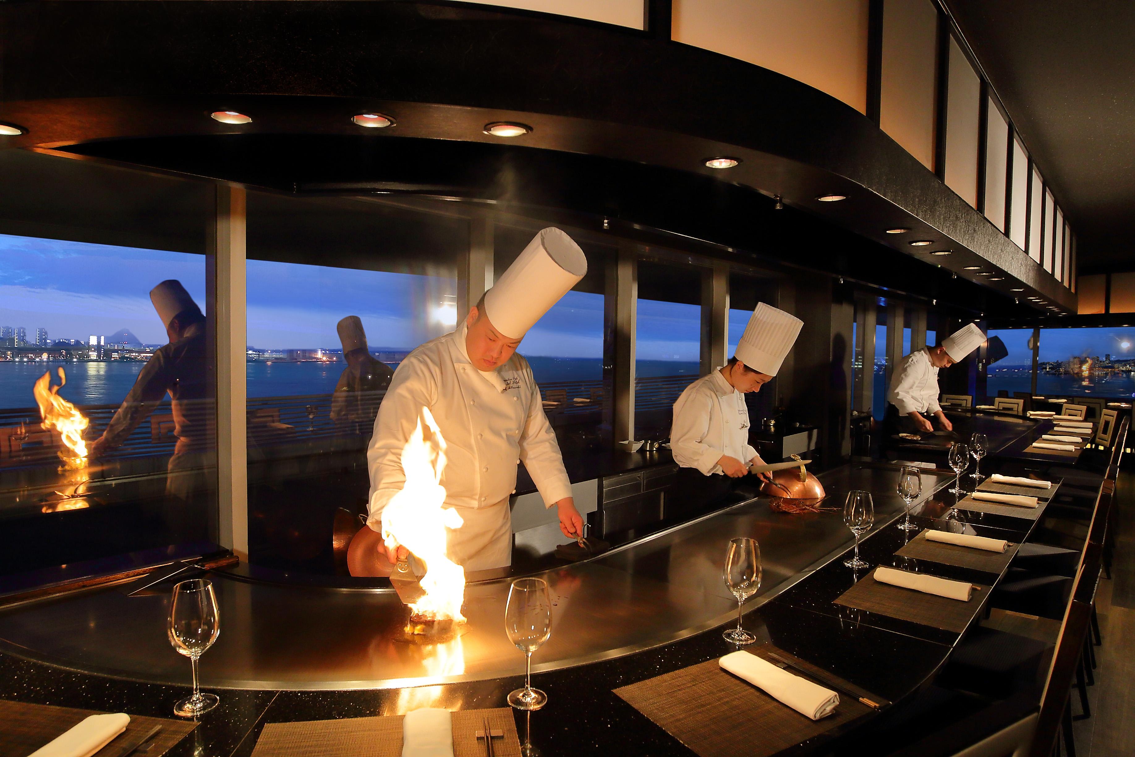 神戸メリケンパークオリエンタルホテル 神戸市 エクステリア 写真 The photo shows a restaurant setting with chefs cooking in an open kitchen, likely in a teppanyaki style. There are three chefs, all wearing white uniforms and tall chef hats, actively engaged in preparing food, with one chef creating a flame on the 