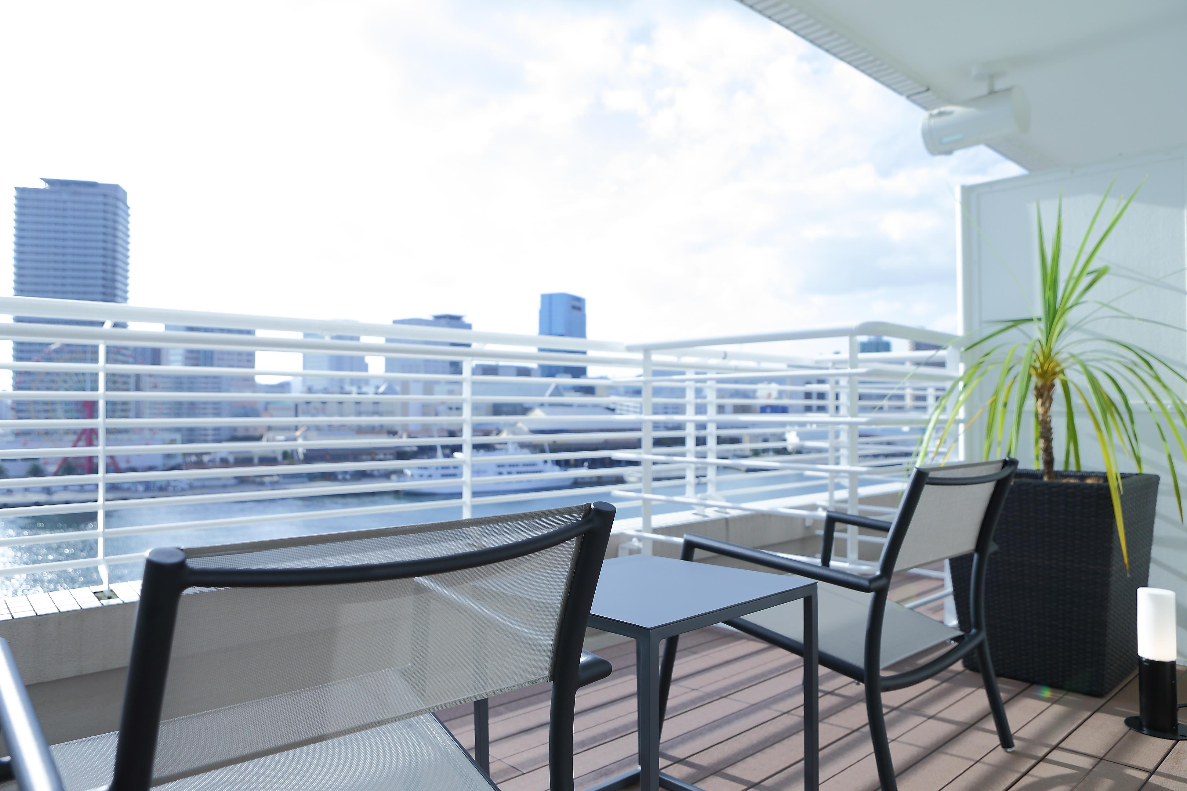 神戸メリケンパークオリエンタルホテル 神戸市 エクステリア 写真 The photo shows a modern balcony or terrace space with a view of a city skyline. There are two black chairs and a small table arranged on a wooden deck. In the background, there are several tall buildings visible, and the sky is partly cloudy. There 