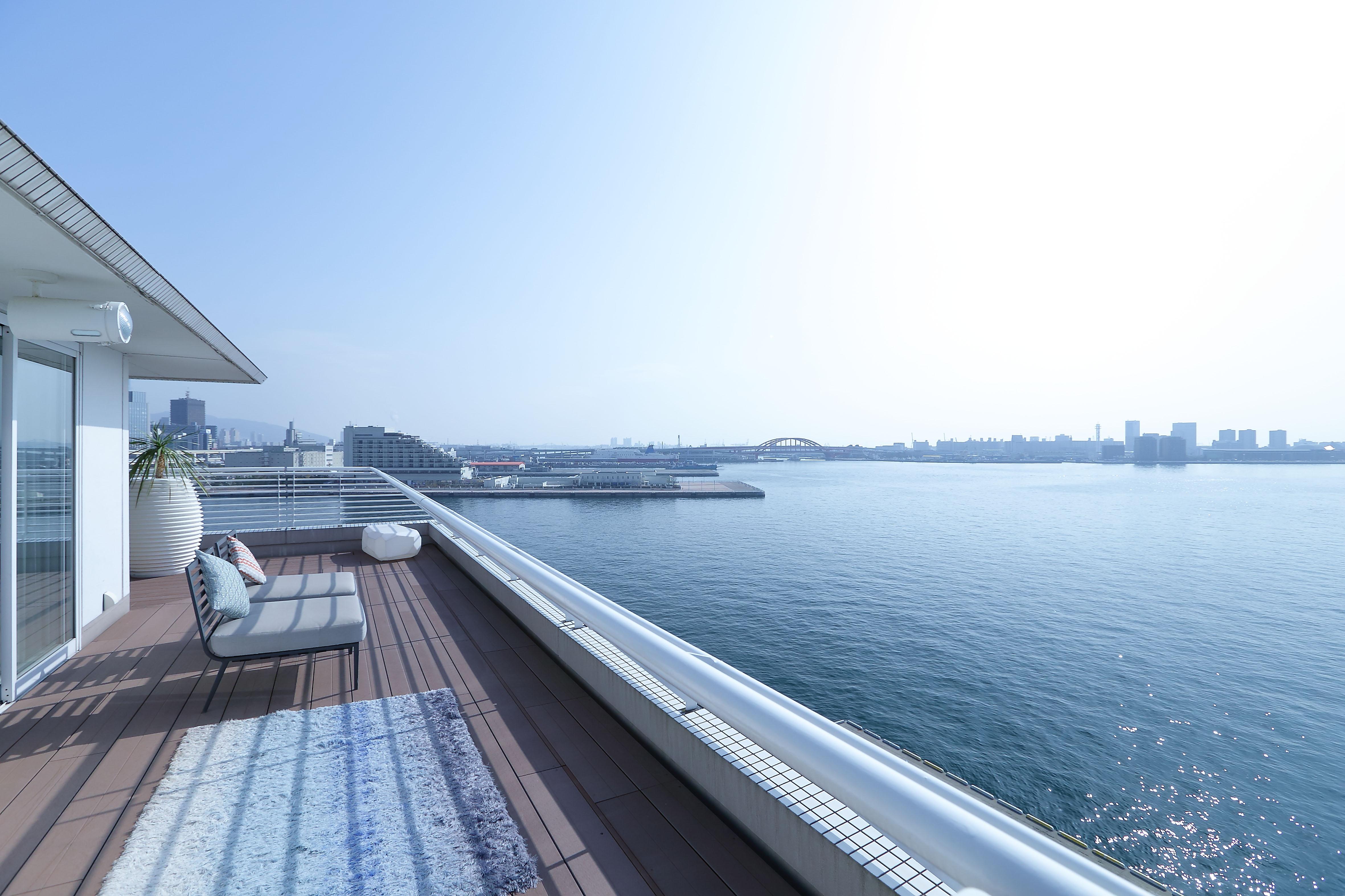 神戸メリケンパークオリエンタルホテル 神戸市 エクステリア 写真 The image shows a spacious balcony or terrace overlooking a body of water, likely a harbor or river, with city buildings in the background. The area is bright and well-lit, indicating a clear, sunny day. There is a modern lounge chair or sofa on the 