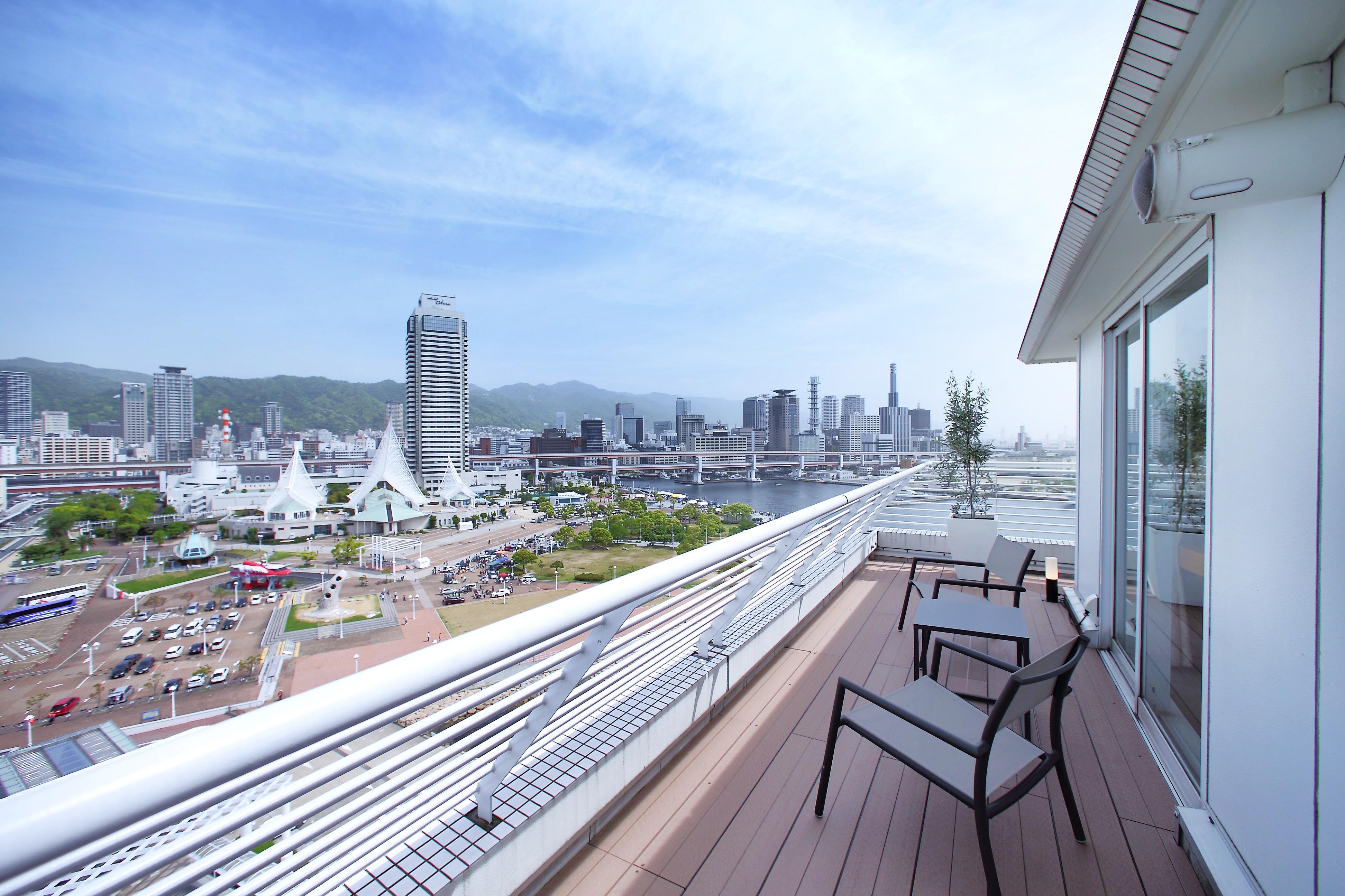 神戸メリケンパークオリエンタルホテル 神戸市 エクステリア 写真 The photo shows a balcony of a modern building with two chairs overlooking a cityscape. In the background, there are several high-rise buildings and a waterfront area. The scene is set against a clear sky with some clouds, and you can see greenery on