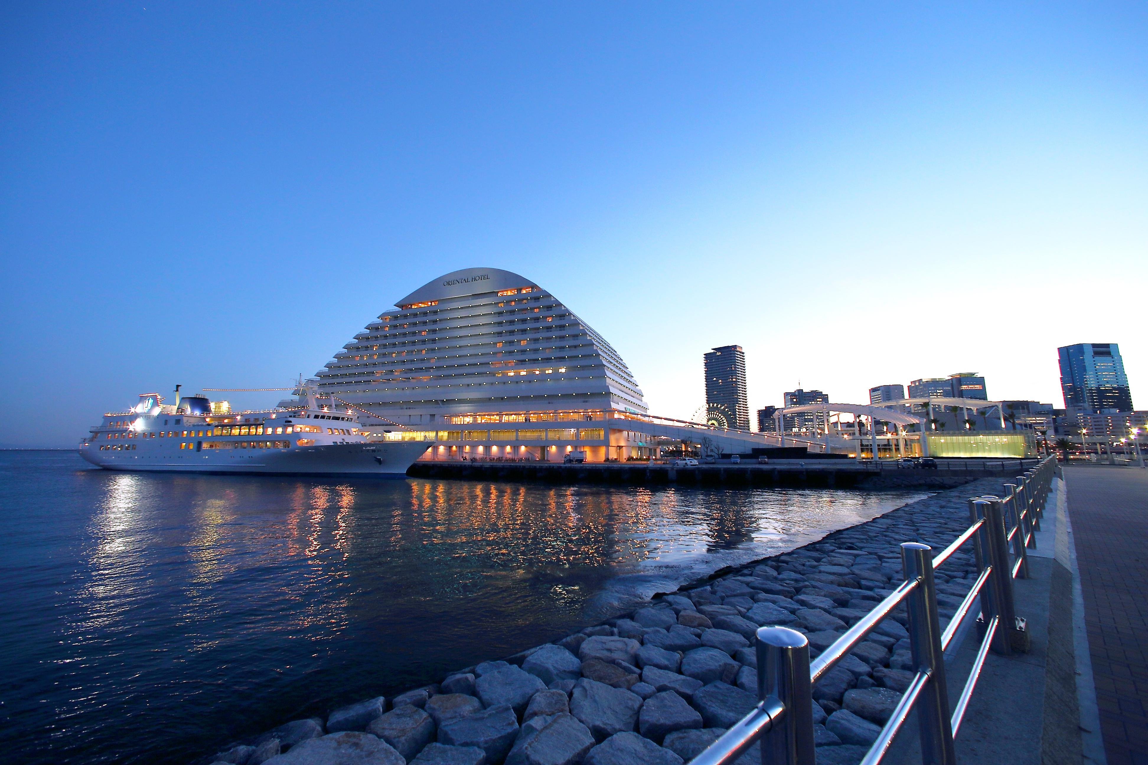 神戸メリケンパークオリエンタルホテル 神戸市 エクステリア 写真 The photo shows a modern building resembling a large, curved structure situated along a waterfront. It is illuminated and reflects beautifully on the calm waters nearby. In the background, there are more buildings typical of an urban skylines, and th