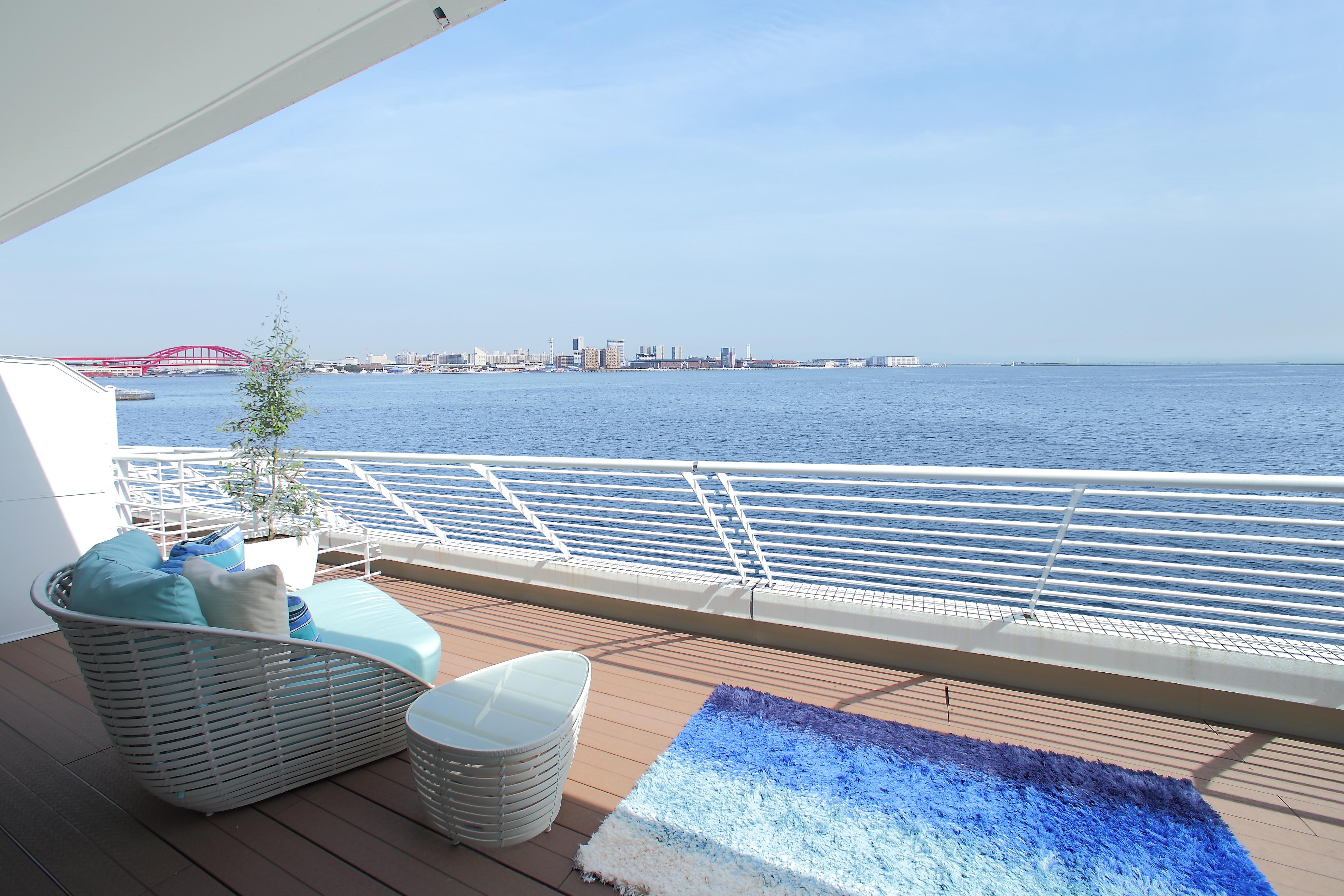 神戸メリケンパークオリエンタルホテル 神戸市 エクステリア 写真 The photo displays a serene outdoor balcony overlooking a body of water. In the foreground, there is a modern, white wicker seating arrangement with blue cushions, accompanied by a small, round table. A soft, gradient rug in shades of blue lies on th