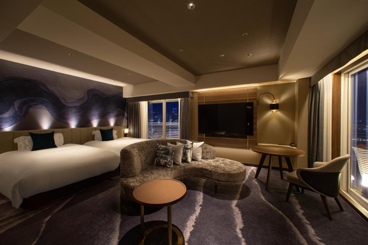 神戸メリケンパークオリエンタルホテル 神戸市 エクステリア 写真 The photo shows a modern hotel room with a stylish interior. There are two beds with white linens, and the decor features warm, ambient lighting. A plush, curved sofa sits in front of a round coffee table, and there’s a chair nearby. A large televisi