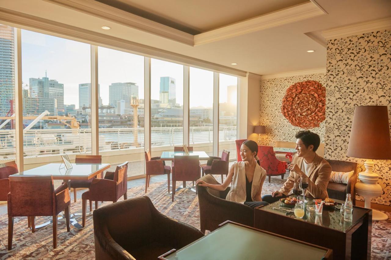 神戸メリケンパークオリエンタルホテル 神戸市 エクステリア 写真 The photo shows a modern, elegantly designed lounge area with large windows providing a view of a cityscape, including buildings by a river. Two people are seated in a cozy setting, enjoying each other's company. The interior features comfortable cha