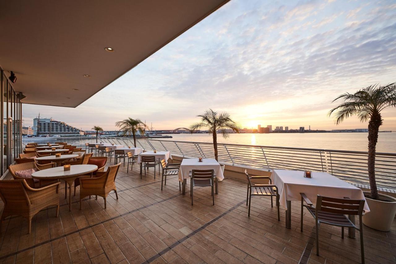 神戸メリケンパークオリエンタルホテル 神戸市 エクステリア 写真 The photo shows a stylish outdoor dining area, likely part of a restaurant or café, situated by the water. There are several tables set up with white tablecloths and wooden chairs. In the background, you can see a beautiful sunset reflecting on the w