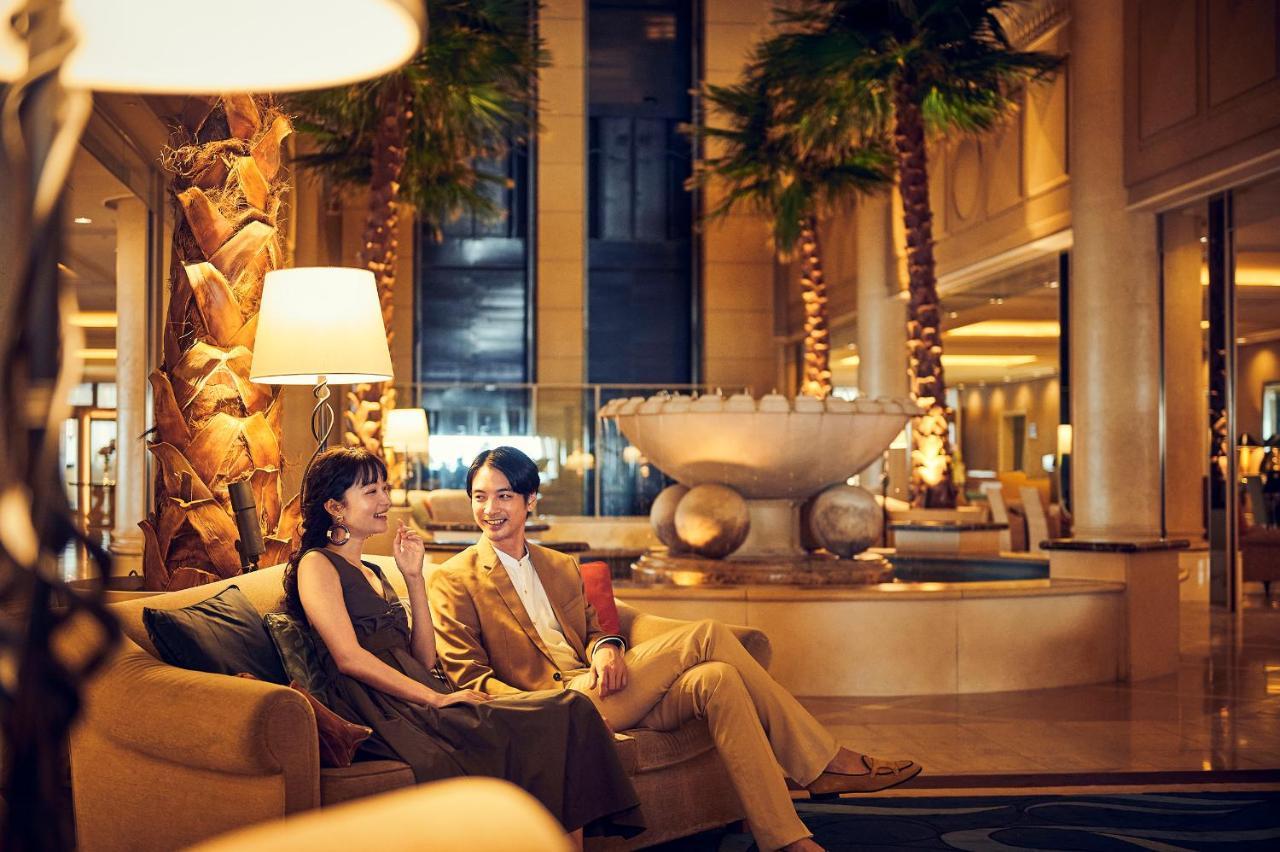 神戸メリケンパークオリエンタルホテル 神戸市 エクステリア 写真 The photo shows two people sitting on a comfortable couch in a luxurious hotel lobby. The setting features elegant decor, including palm trees and a large decorative fountain in the background. The individuals appear to be having a pleasant conversat