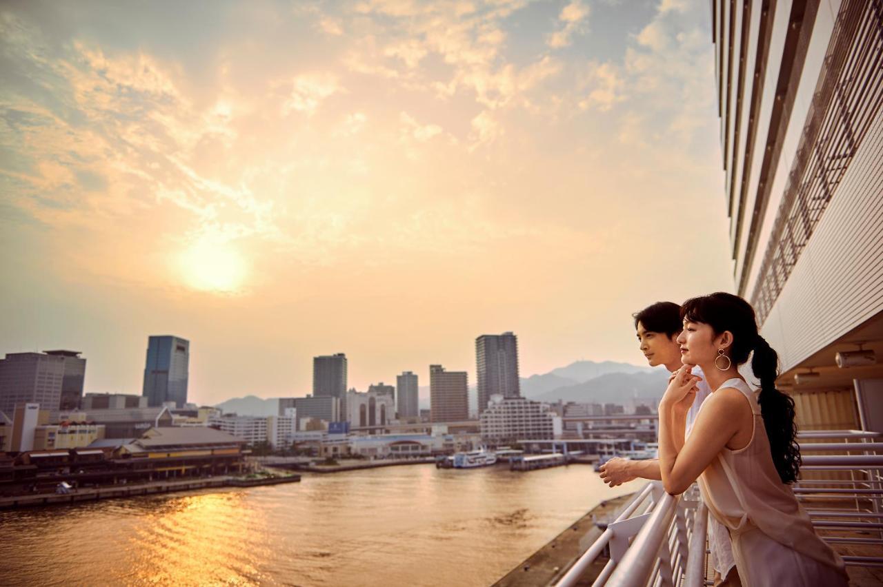 神戸メリケンパークオリエンタルホテル 神戸市 エクステリア 写真 The photo depicts two people standing on a balcony, gazing out over a cityscape illuminated by a warm sunset. The scene captures a river reflecting the golden hues of the sky, with various buildings lining the waterfront. In the background, mountains