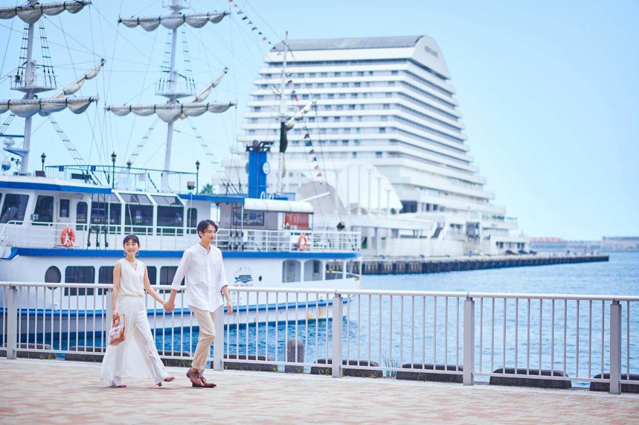 神戸メリケンパークオリエンタルホテル 神戸市 エクステリア 写真 The photo showcases a couple walking hand-in-hand along a waterfront promenade. They are dressed in light, elegant clothing suitable for warm weather. Behind them, there are boats docked, including a large ship and a two-masted vessel. The background