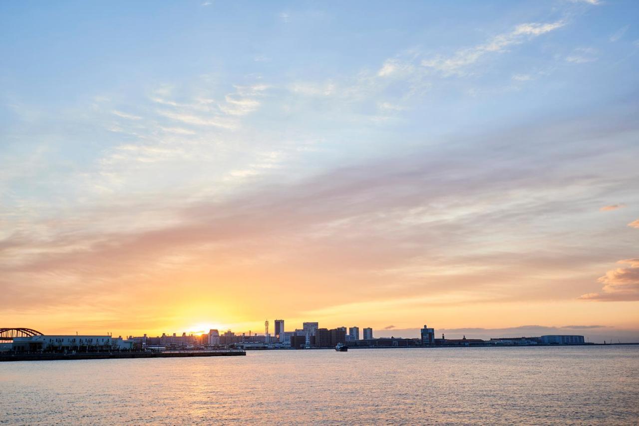 神戸メリケンパークオリエンタルホテル 神戸市 エクステリア 写真 The photo shows a serene sunset over a body of water, likely an ocean or bay. The sky is painted in soft hues of orange, pink, and blue, creating a tranquil atmosphere. In the foreground, calm water reflects the colors of the sky. In the background, 