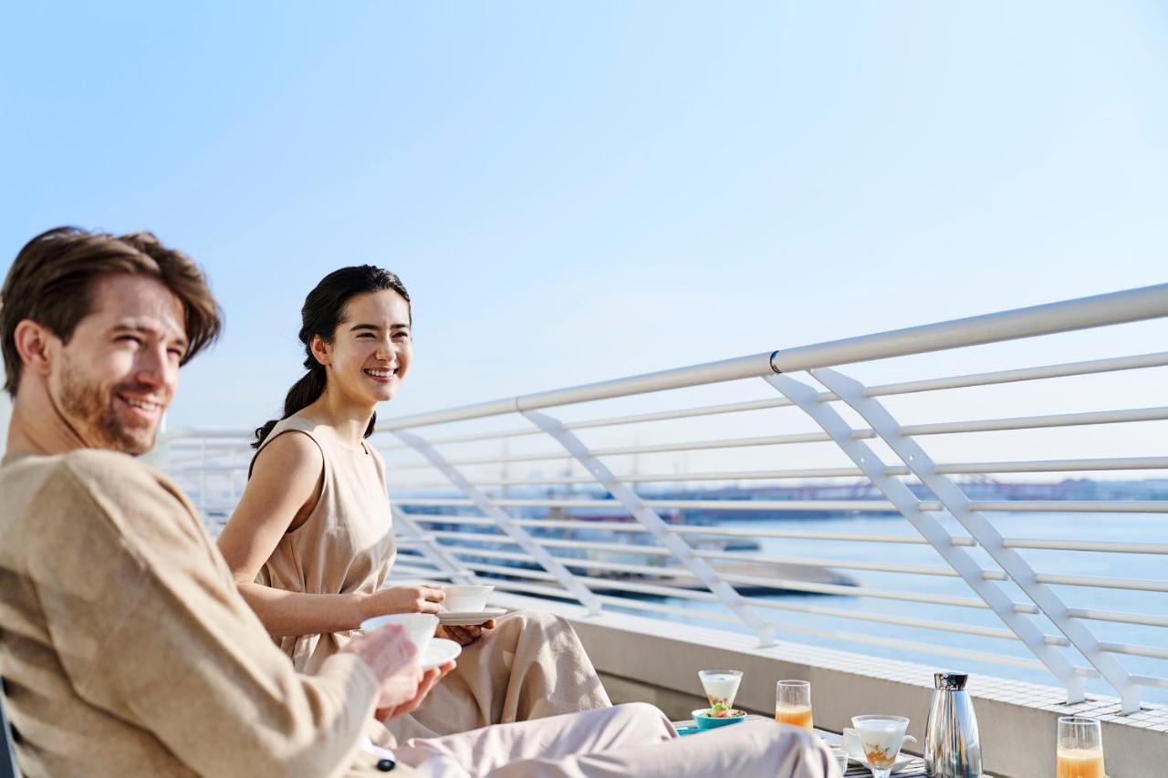 神戸メリケンパークオリエンタルホテル 神戸市 エクステリア 写真 The photo shows two people enjoying a pleasant moment outdoors, seated on a balcony or terrace. The scene has a bright, sunny atmosphere with blue skies and water in the background. The man has a casual look with a light-colored outfit, while the wom
