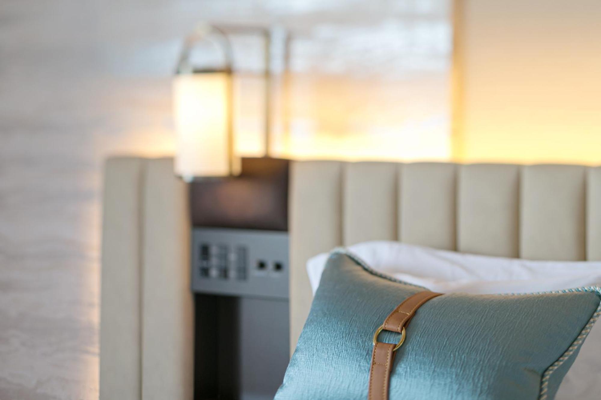 神戸メリケンパークオリエンタルホテル 神戸市 エクステリア 写真 The photo shows a cozy and stylish bedroom setting. In the foreground, there is a decorative pillow with a blue fabric and a leather accent, resting on a beige upholstered bed. Behind the bed, there is a softly lit ambiance with a lamp or wall sconce