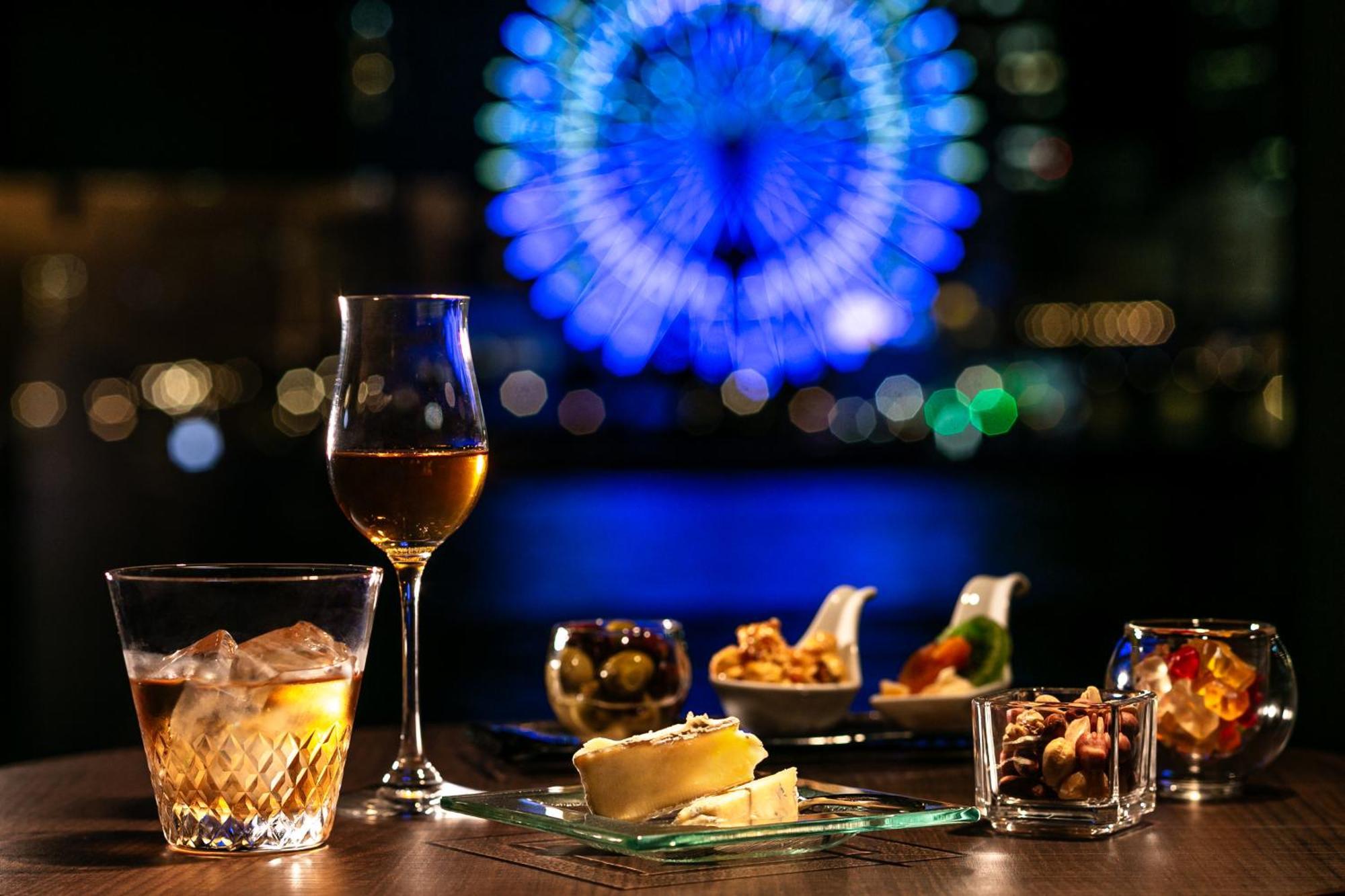 神戸メリケンパークオリエンタルホテル 神戸市 エクステリア 写真 The photo shows a dining setup with a variety of appetizers and drinks arranged on a table. In the foreground, there is a glass of clear liquid with ice and a wine glass filled with a golden beverage. A platter features several small dishes containin