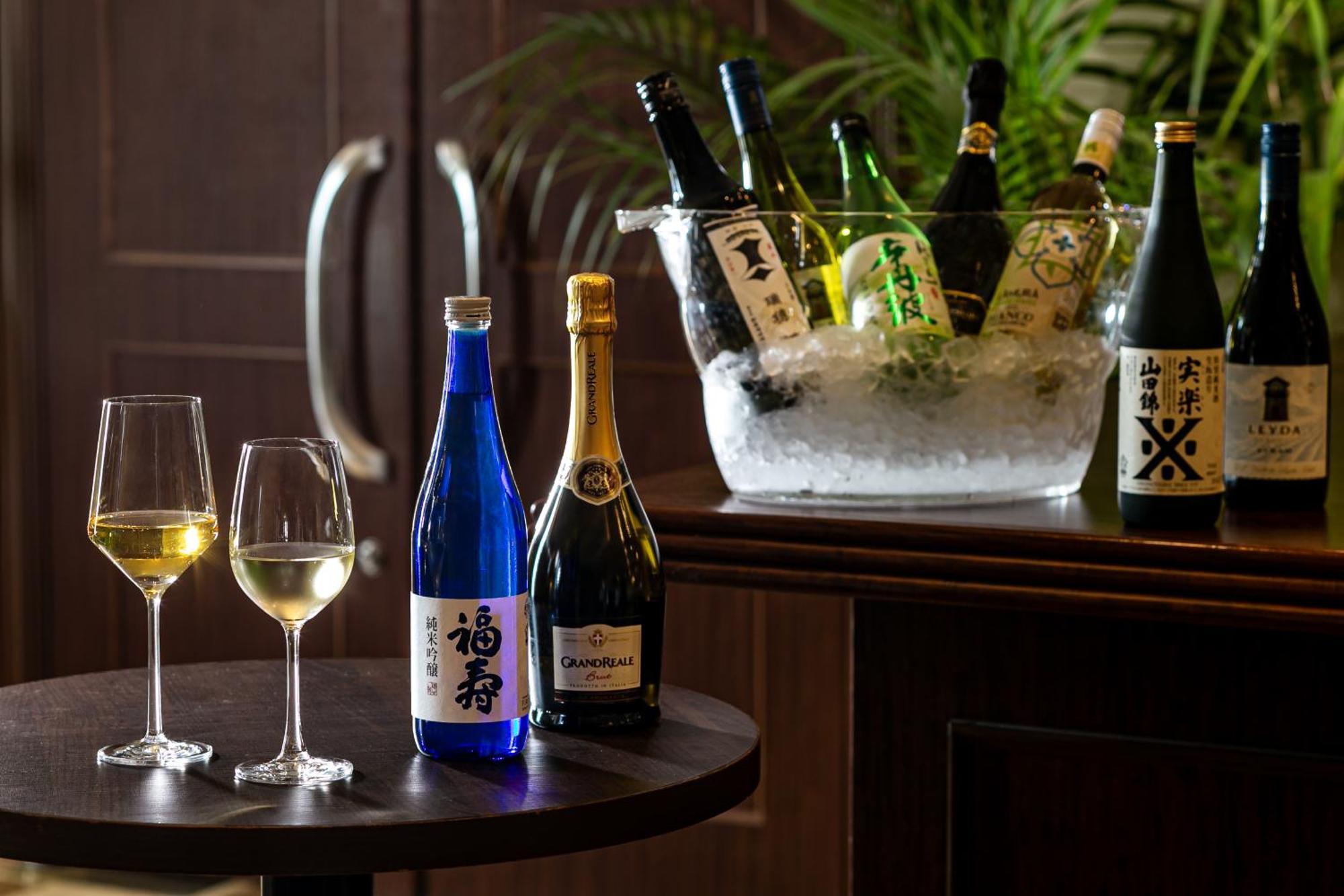神戸メリケンパークオリエンタルホテル 神戸市 エクステリア 写真 The photo features a selection of alcoholic beverages displayed elegantly. On a dark wooden table, there are two wine glasses filled with a light-colored liquid, possibly white wine. Nearby, there is a blue bottle labeled with Japanese characters, al