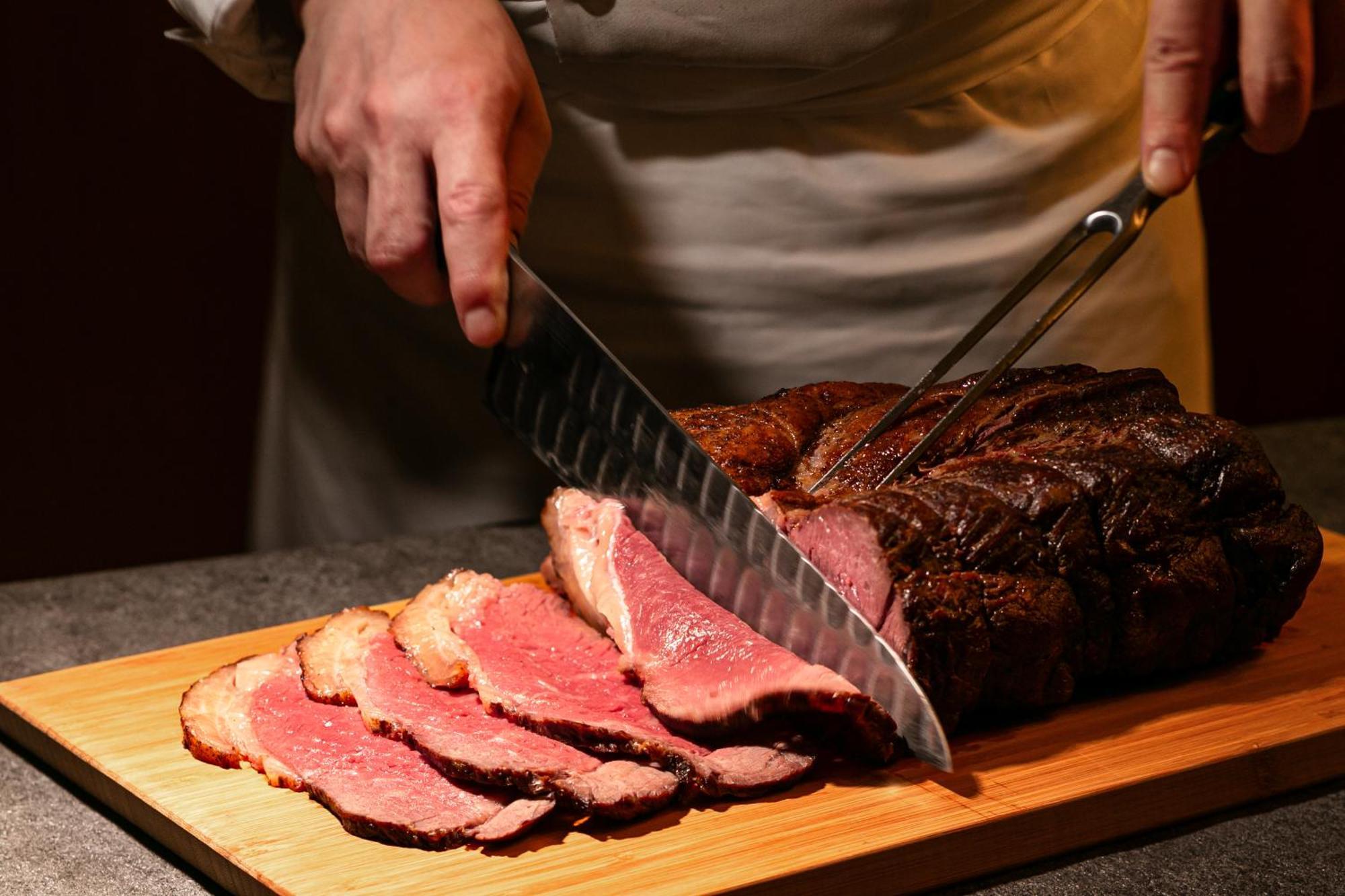 神戸メリケンパークオリエンタルホテル 神戸市 エクステリア 写真 The photo shows a person slicing a large piece of meat, likely a roast or steak, on a wooden cutting board. The meat is beautifully cooked, with pink slices being cut and arranged. The person is using a sharp knife and fork, focused on the task of ca