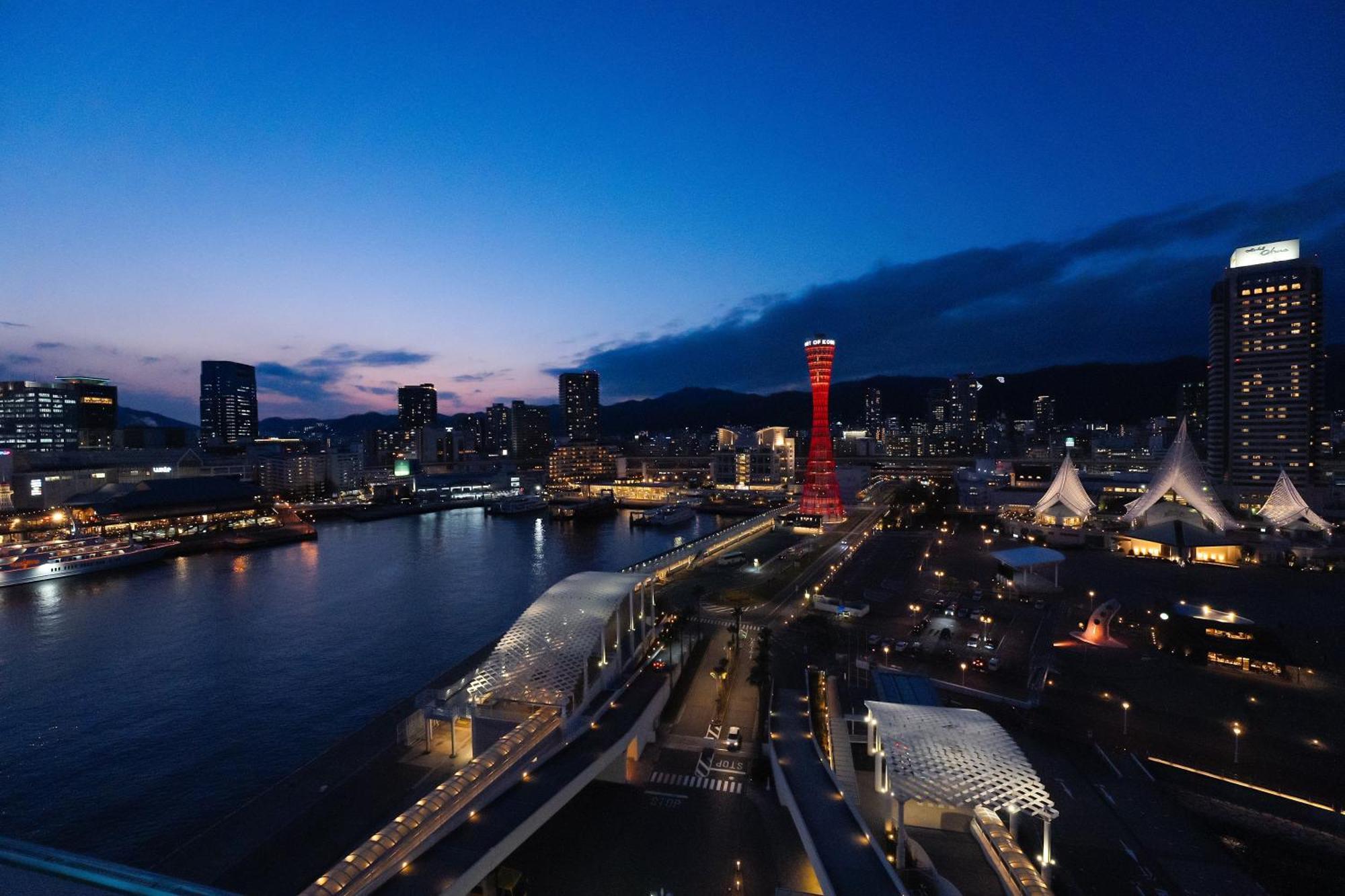 神戸メリケンパークオリエンタルホテル 神戸市 エクステリア 写真 The photo depicts a scenic view of a waterfront city at dusk. The sky transitions from blue to darker shades as night falls. A prominent red-lit tower stands out against the skyline, surrounded by modern buildings. The waterfront features a bridge an