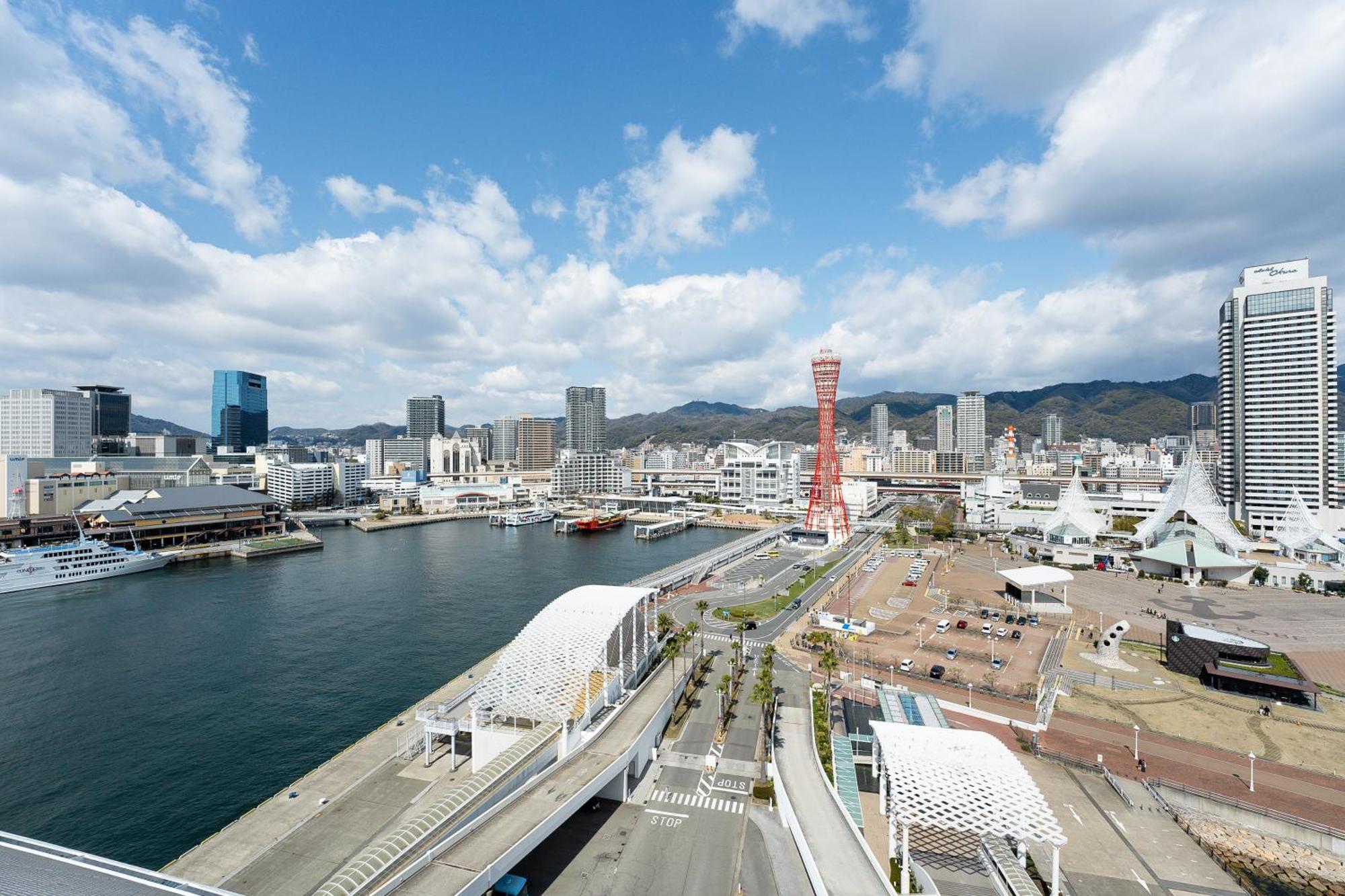 神戸メリケンパークオリエンタルホテル 神戸市 エクステリア 写真 The photo shows a coastal cityscape, likely featuring a harbor or waterfront area. There is a mix of modern buildings, some of which are tall skyscrapers, along with a notable tower prominently visible, possibly indicating a landmark. 

In the foregr