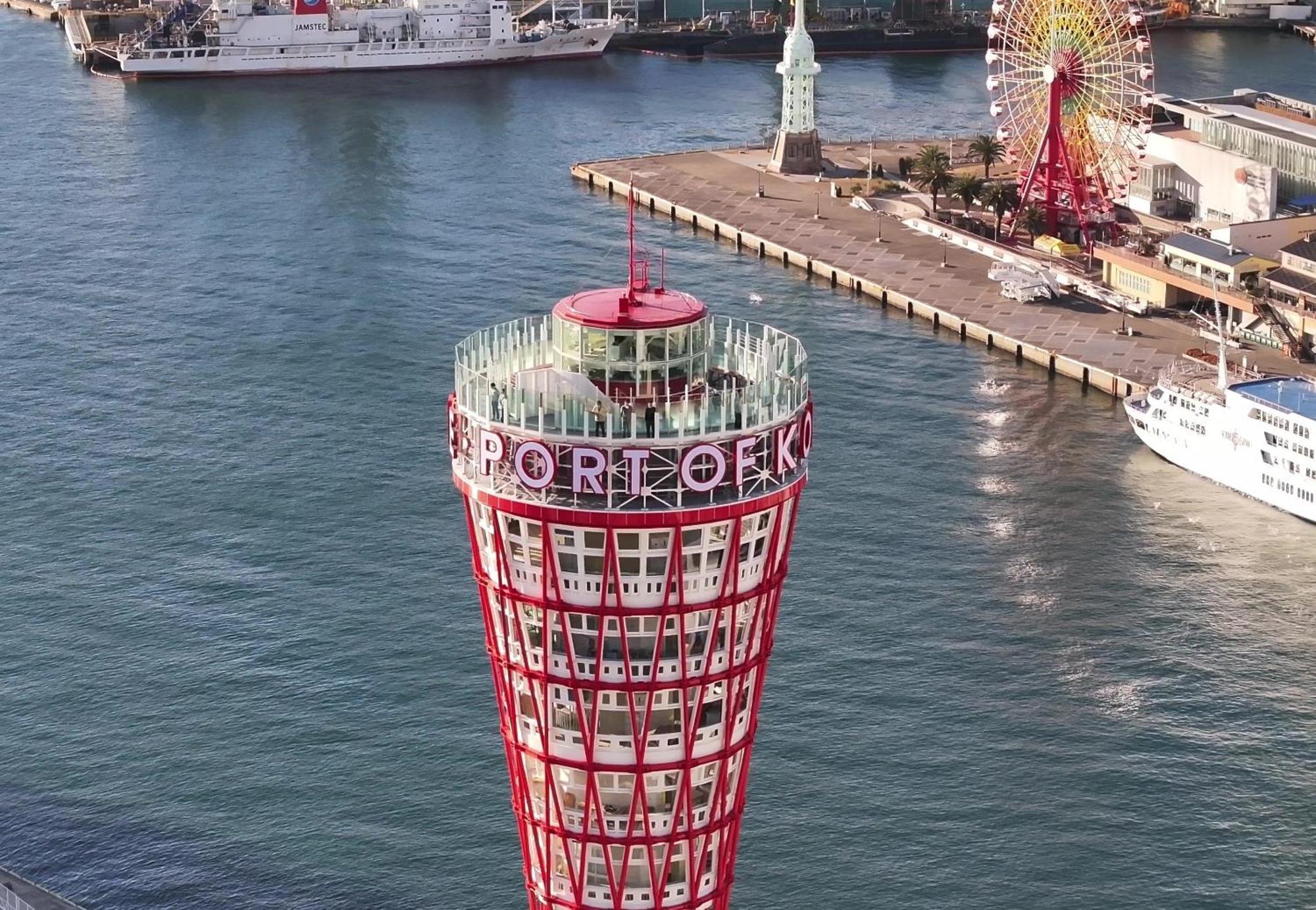 神戸メリケンパークオリエンタルホテル 神戸市 エクステリア 写真 The photo shows a prominent tower with a unique, modern design. The tower is cone-shaped and colored in red and white, with the words "PORT OF KOBE" displayed prominently on it. Surrounding the tower, there are boats in the water, and a ferris wheel 