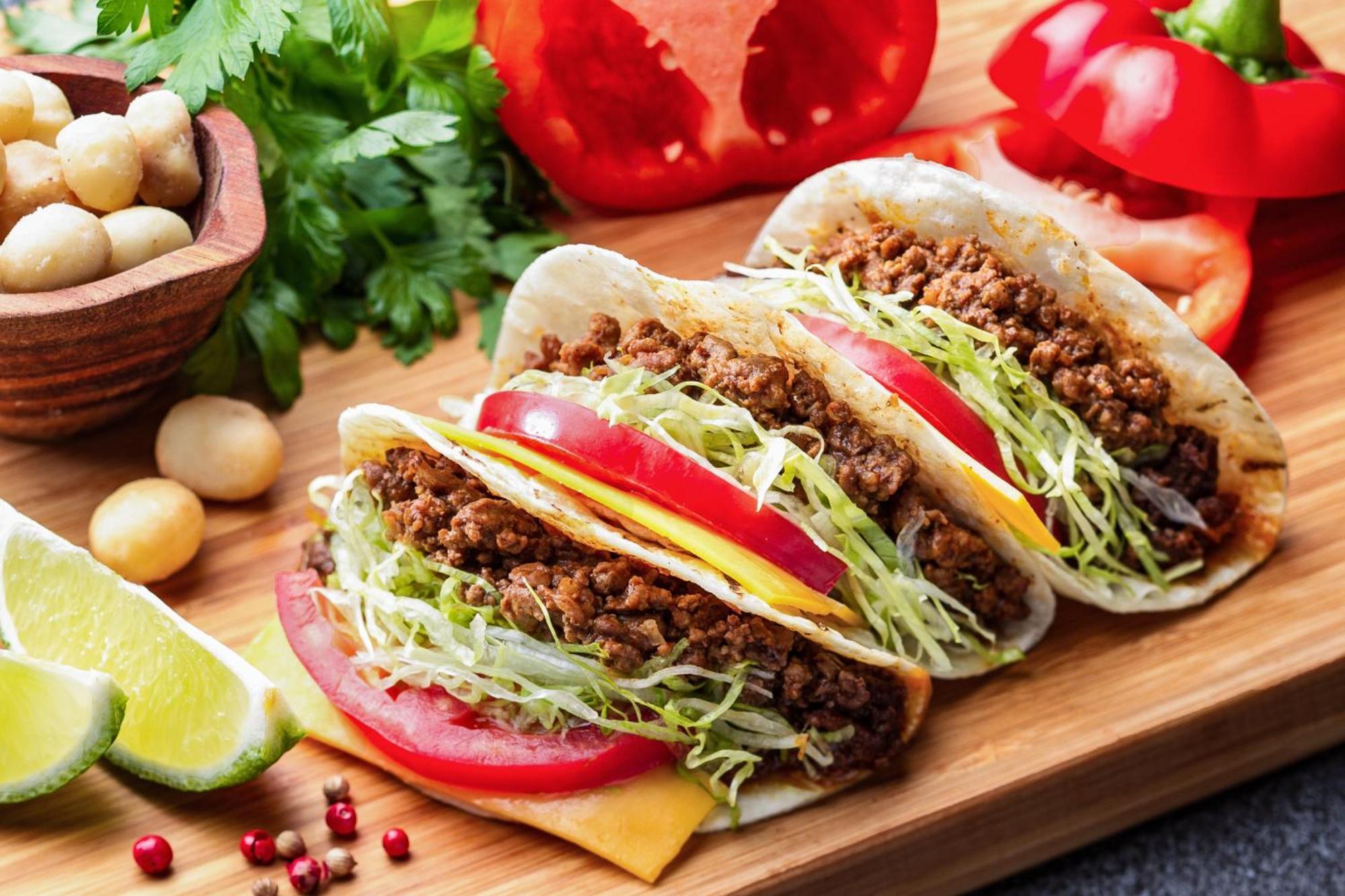 神戸メリケンパークオリエンタルホテル 神戸市 エクステリア 写真 The photo shows three tacos placed on a wooden serving board. Each taco is filled with seasoned ground meat, topped with shredded lettuce and slices of tomato. There are also slices of cheese visible inside the tacos. Surrounding the tacos are fresh 