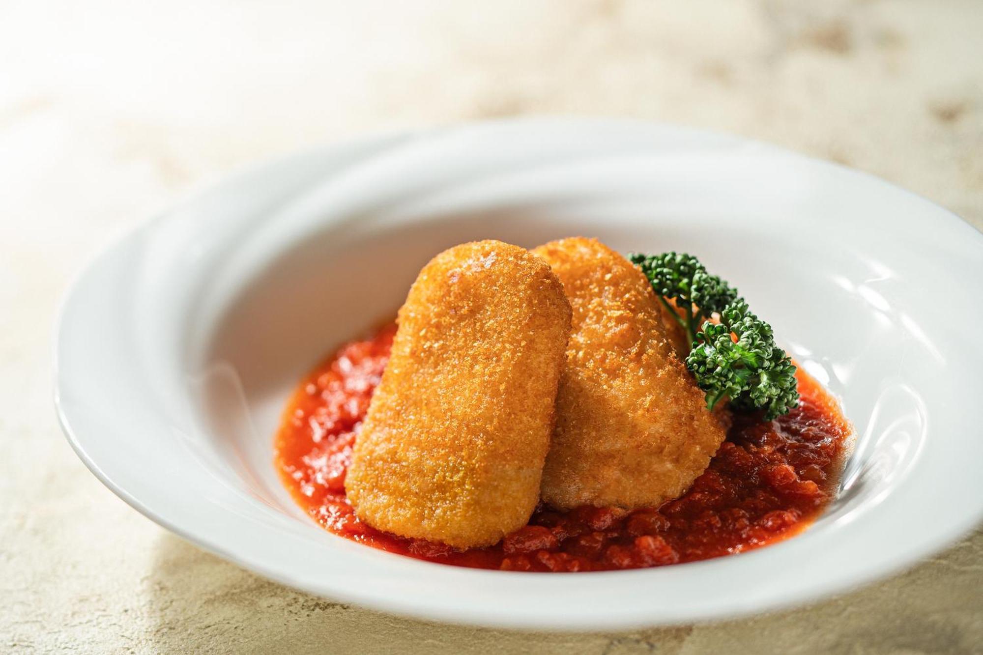 神戸メリケンパークオリエンタルホテル 神戸市 エクステリア 写真 The photo shows two breaded and fried pieces of food, likely croquettes or a similar dish, placed on a bed of rich red tomato sauce. There is a small garnish of green broccoli beside the croquettes, adding a touch of color to the presentation. The di