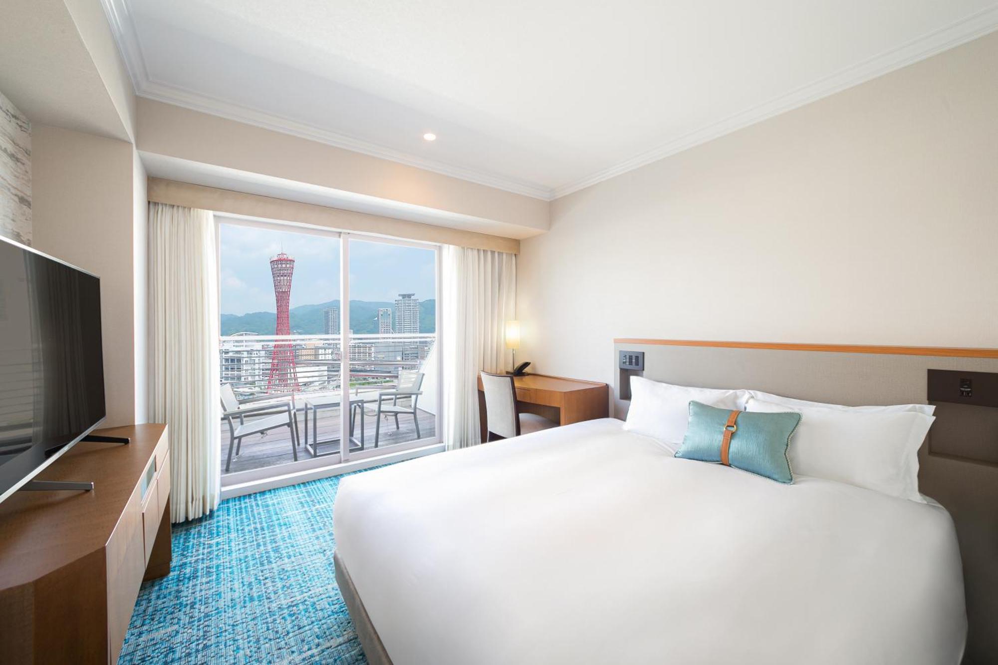 神戸メリケンパークオリエンタルホテル 神戸市 エクステリア 写真 The photo shows a modern hotel room with a large bed featuring white bedding and a teal accent pillow. There's a wooden desk and a flat-screen TV opposite the bed. Large windows provide a view of an outdoor area, which includes a balcony with furnitu