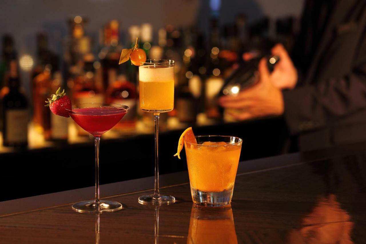 神戸メリケンパークオリエンタルホテル 神戸市 エクステリア 写真 The photo displays three cocktails arranged on a bar. 

1. **Left**: A vibrant pink cocktail served in a tall, slender glass, garnished with a fresh strawberry.
2. **Middle**: A light yellow drink in a frosted glass with a citrus twist, topped with a