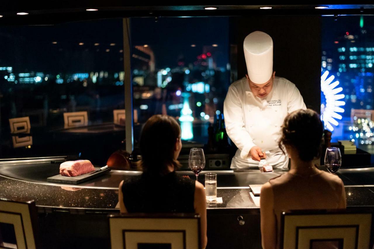 神戸メリケンパークオリエンタルホテル 神戸市 エクステリア 写真 The photo depicts a dining scene in an upscale restaurant. In the foreground, two patrons are seated at a counter, watching a chef who is preparing food. The chef, wearing a traditional white chef's hat and coat, is engaged in the cooking process, li