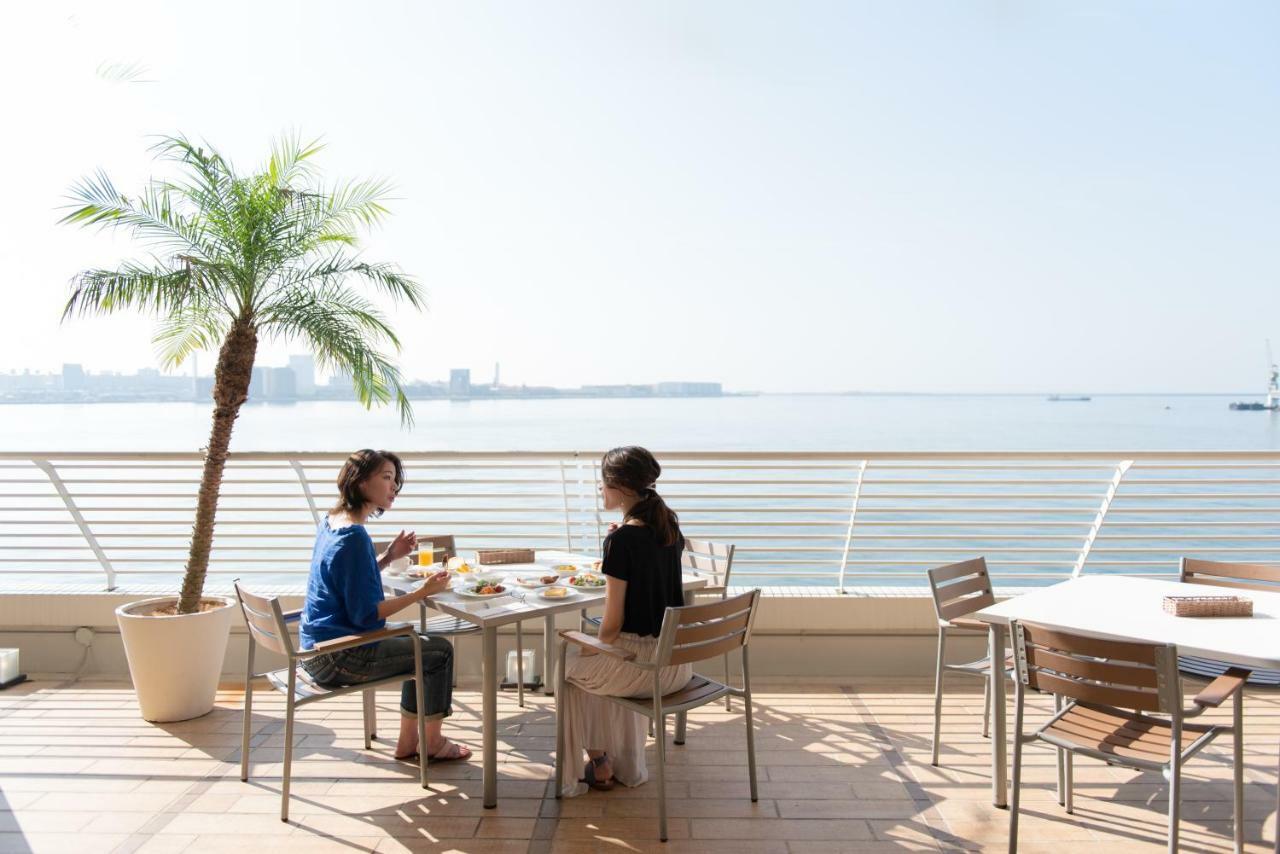 神戸メリケンパークオリエンタルホテル 神戸市 エクステリア 写真 The photo shows two women sitting at a table on a terrace overlooking a body of water. They appear to be enjoying a meal together. The setting has a tropical vibe, with a small palm tree in a pot nearby and clear skies visible in the background. The 