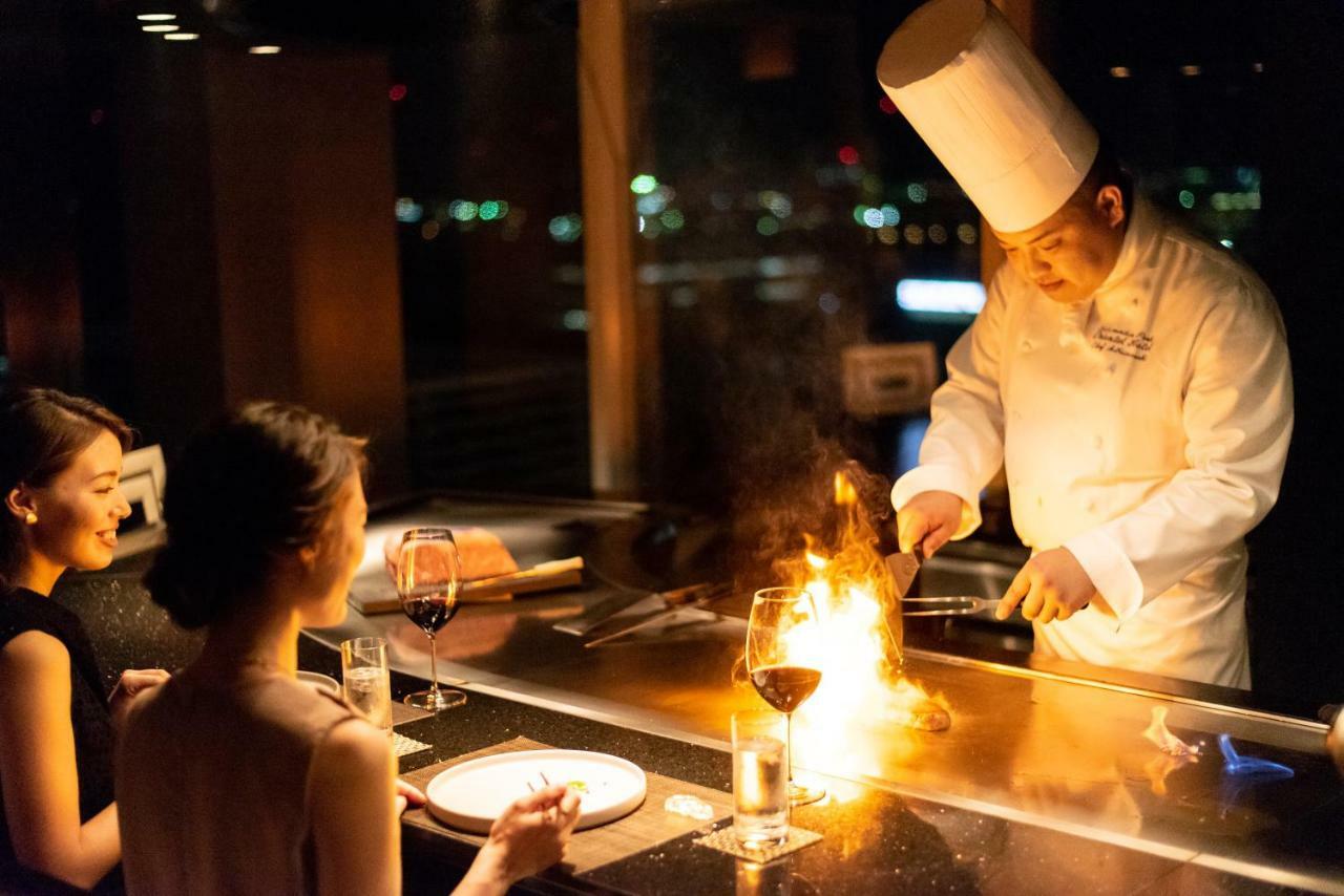 神戸メリケンパークオリエンタルホテル 神戸市 エクステリア 写真 The photo shows a chef performing a cooking demonstration at a teppanyaki restaurant. The chef is wearing a traditional white chef's hat and jacket, and is cooking in front of two diners. One of the cooking techniques involves flames, creating a dram