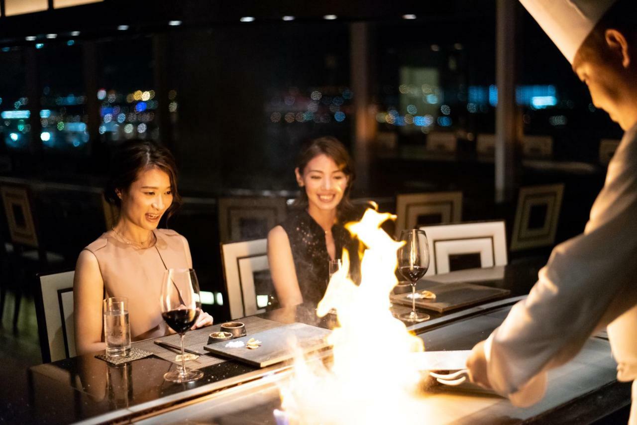神戸メリケンパークオリエンタルホテル 神戸市 エクステリア 写真 The photo depicts a dining scene in a luxurious restaurant. Two women are seated at a table, enjoying the experience. They appear to be engaged and delighted by the culinary performance taking place in front of them. A chef is preparing food on a gri