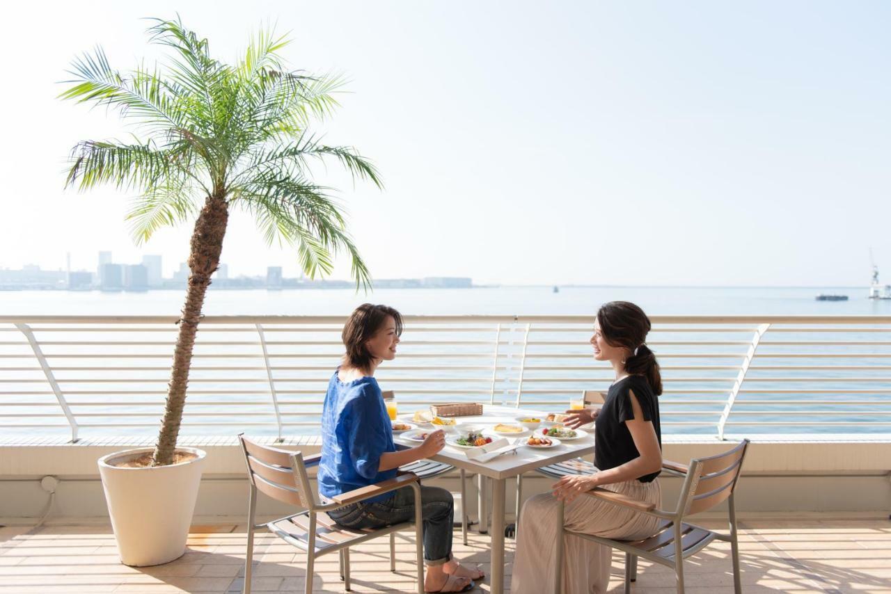 神戸メリケンパークオリエンタルホテル 神戸市 エクステリア 写真 The photo shows two women sitting at a table on a sunny terrace by the water. They are enjoying a meal together, with dishes laid out on the table in front of them. There is a tall palm tree in a planter nearby, and the view overlooks a body of water
