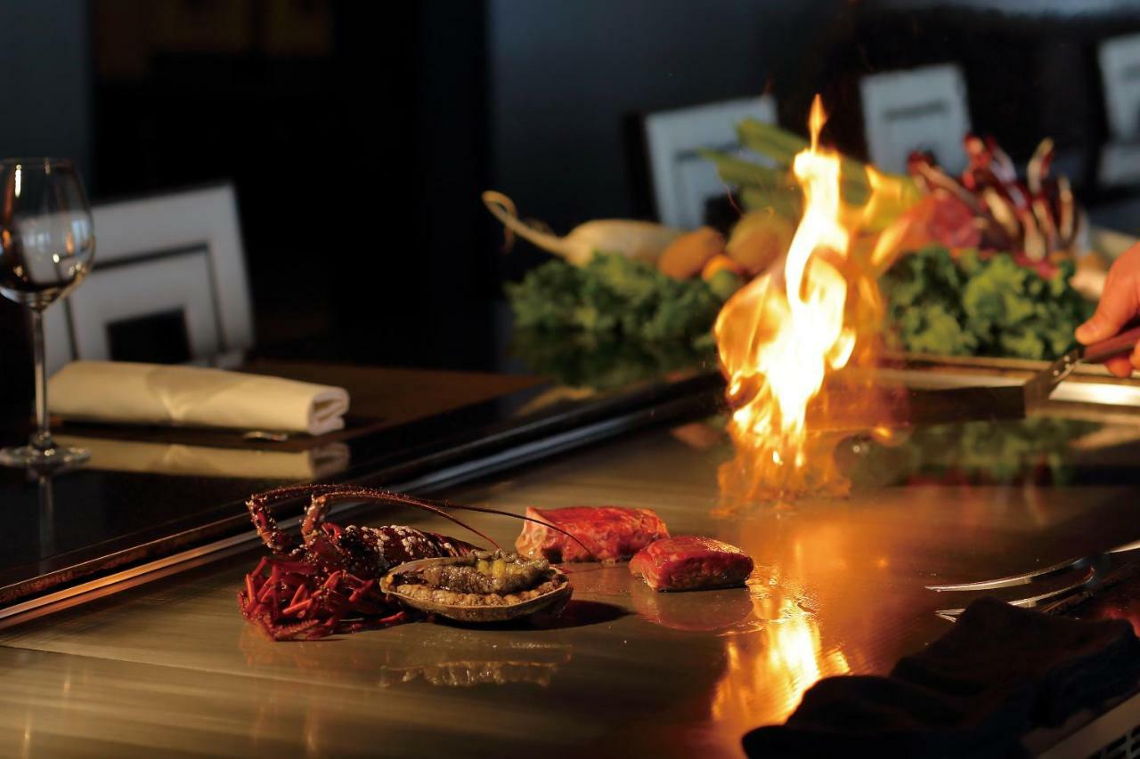 神戸メリケンパークオリエンタルホテル 神戸市 エクステリア 写真 The photo depicts a cooking scene, likely at a teppanyaki grill or similar style of dining. There are pieces of meat, including what appears to be steak, laid out on a shiny metal cooking surface. In the foreground, there is a flame rising from the g