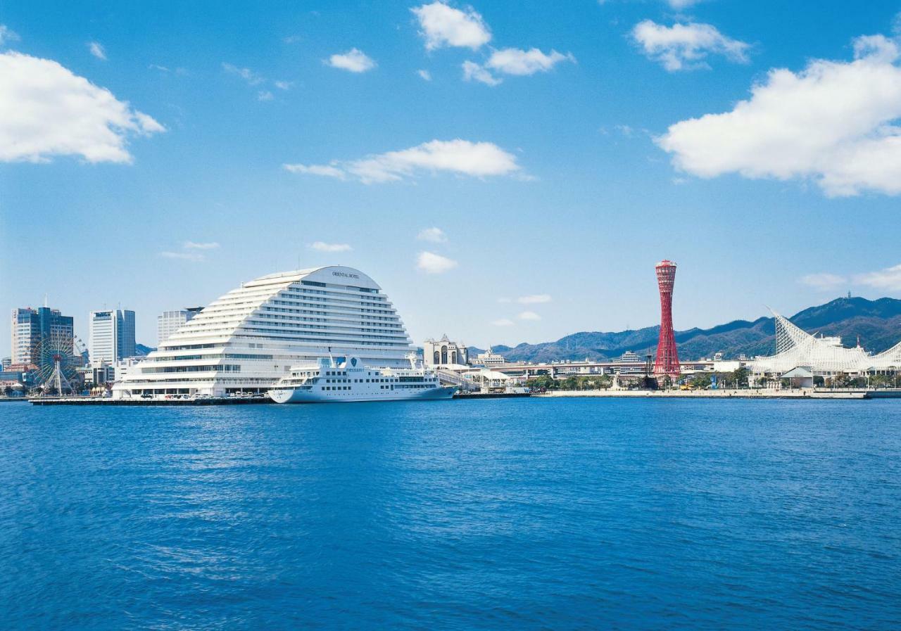 神戸メリケンパークオリエンタルホテル 神戸市 エクステリア 写真 The photo depicts a coastal scene featuring a large, modern building with a distinctive curved design situated near the water. This building is likely a hotel or commercial facility. Next to it, there is a tall, red observation tower, which could ser