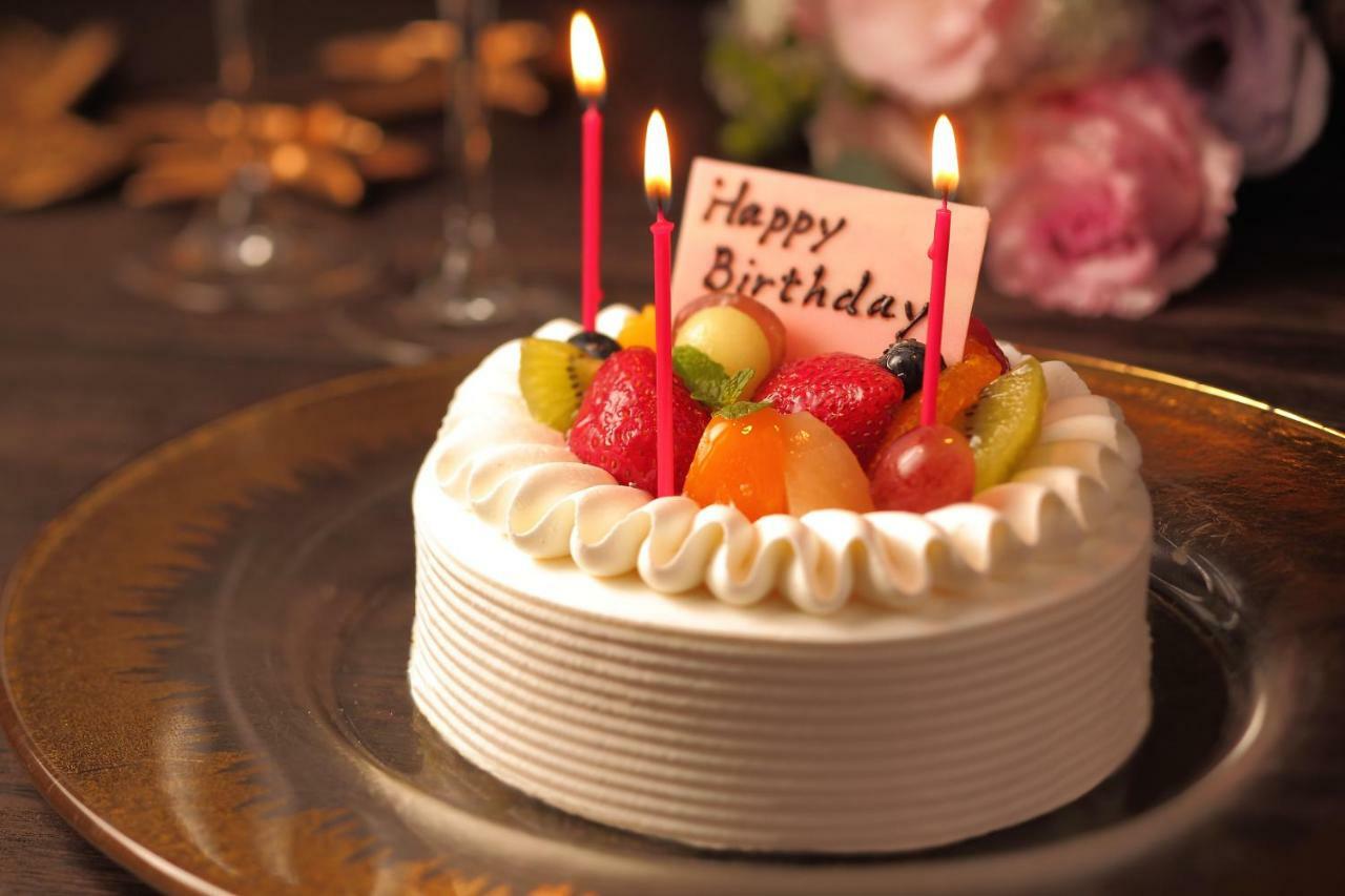 神戸メリケンパークオリエンタルホテル 神戸市 エクステリア 写真 The photo shows a beautifully decorated birthday cake placed on a plate. The cake is round with white frosting, and it features a variety of fresh fruits on top, including strawberries, grapes, and kiwis. There are four red candles lit on the cake, a