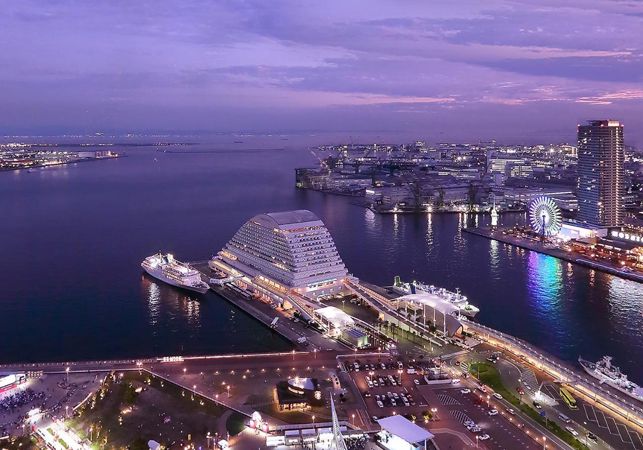 神戸メリケンパークオリエンタルホテル 神戸市 エクステリア 写真 The photo depicts a scenic view of a harbor at dusk. The scene includes modern architectural structures along the waterfront, with one building having a unique, curved design. There are several boats docked in the water, and a colorful Ferris wheel i